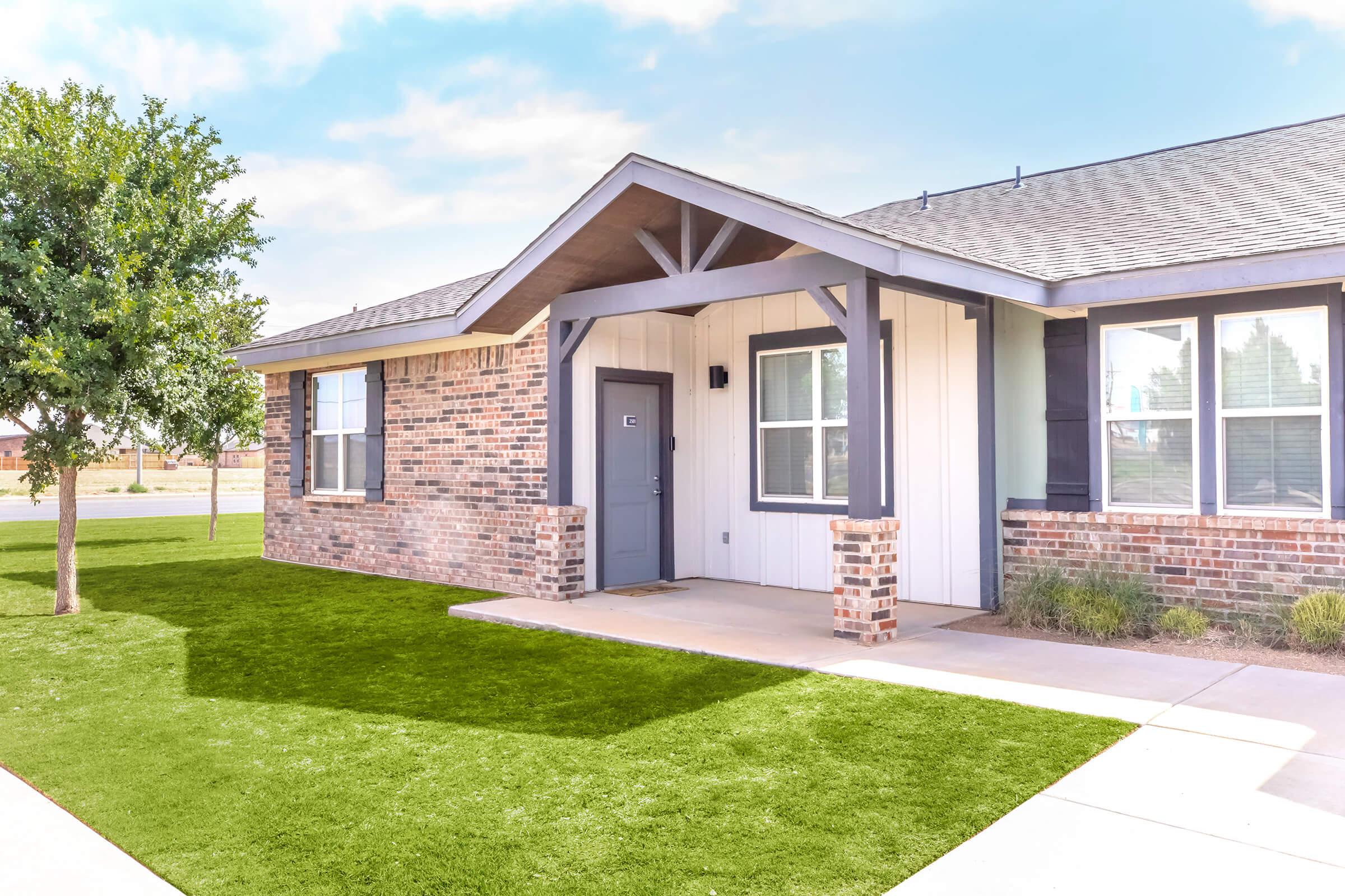 A modern single-story home featuring a combination of brick and siding exterior, a welcoming front porch with a gray door, and neatly maintained landscaping. The green lawn contrasts with the home’s neutral colors, under a clear blue sky.