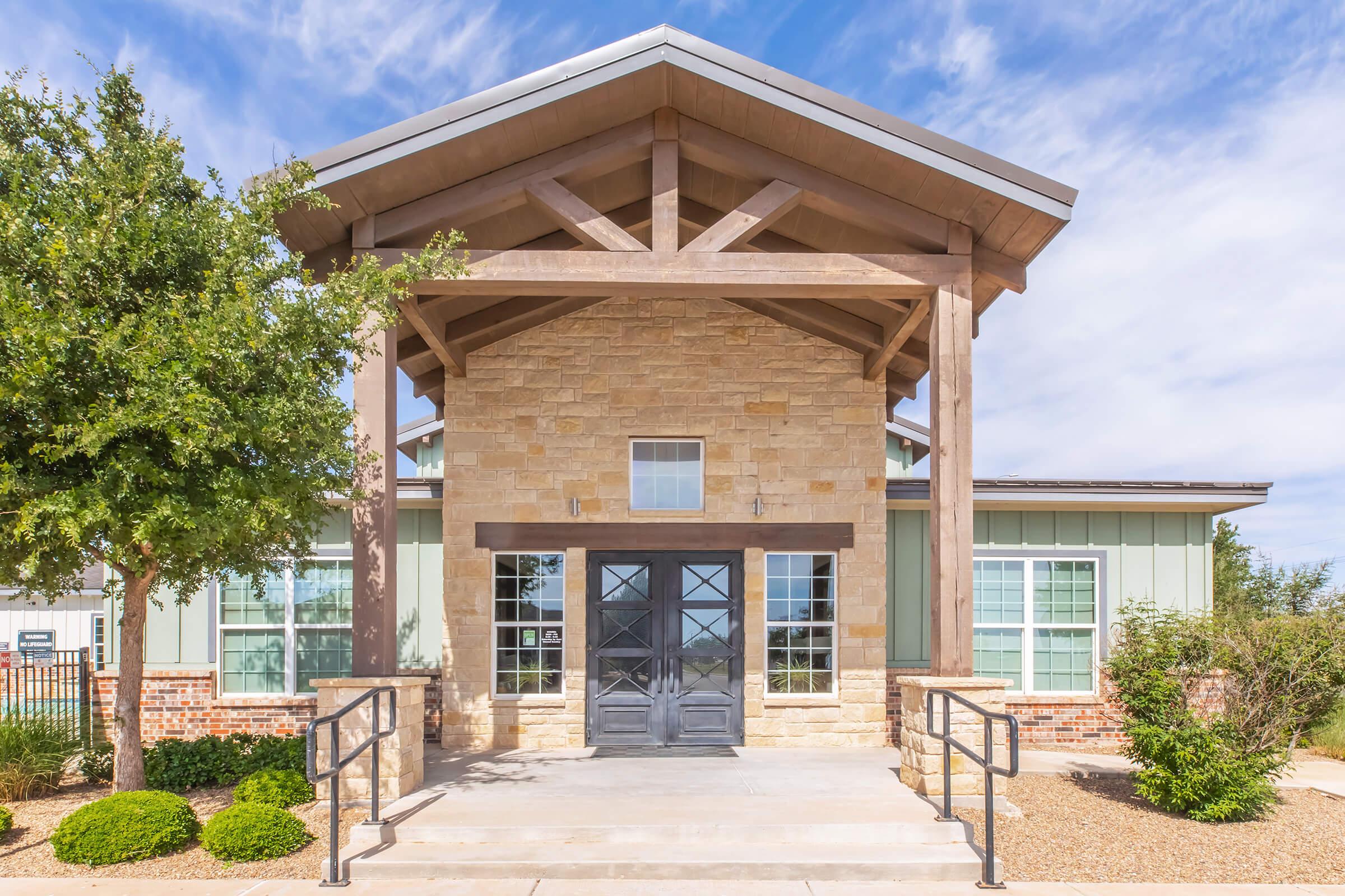 A modern building with a large wooden entrance, featuring stone and siding materials. The facade includes large windows and a peaked roof supported by wooden beams. Surrounding the entrance are landscaped elements like shrubs, with a clear blue sky above.