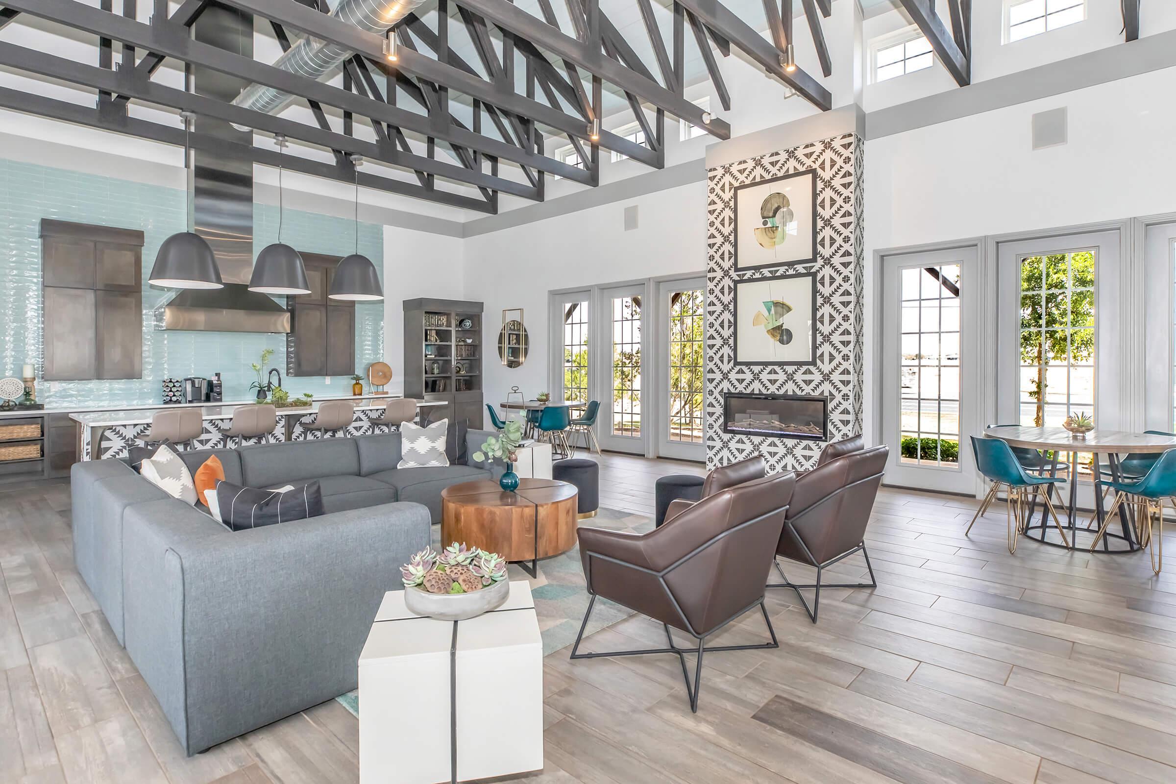 A spacious and modern living area featuring a comfortable gray sofa and stylish brown chairs. The room has high wooden beams, large windows for natural light, and a statement wall with decorative patterns. A dining table is visible in the background, along with a well-equipped kitchen area.