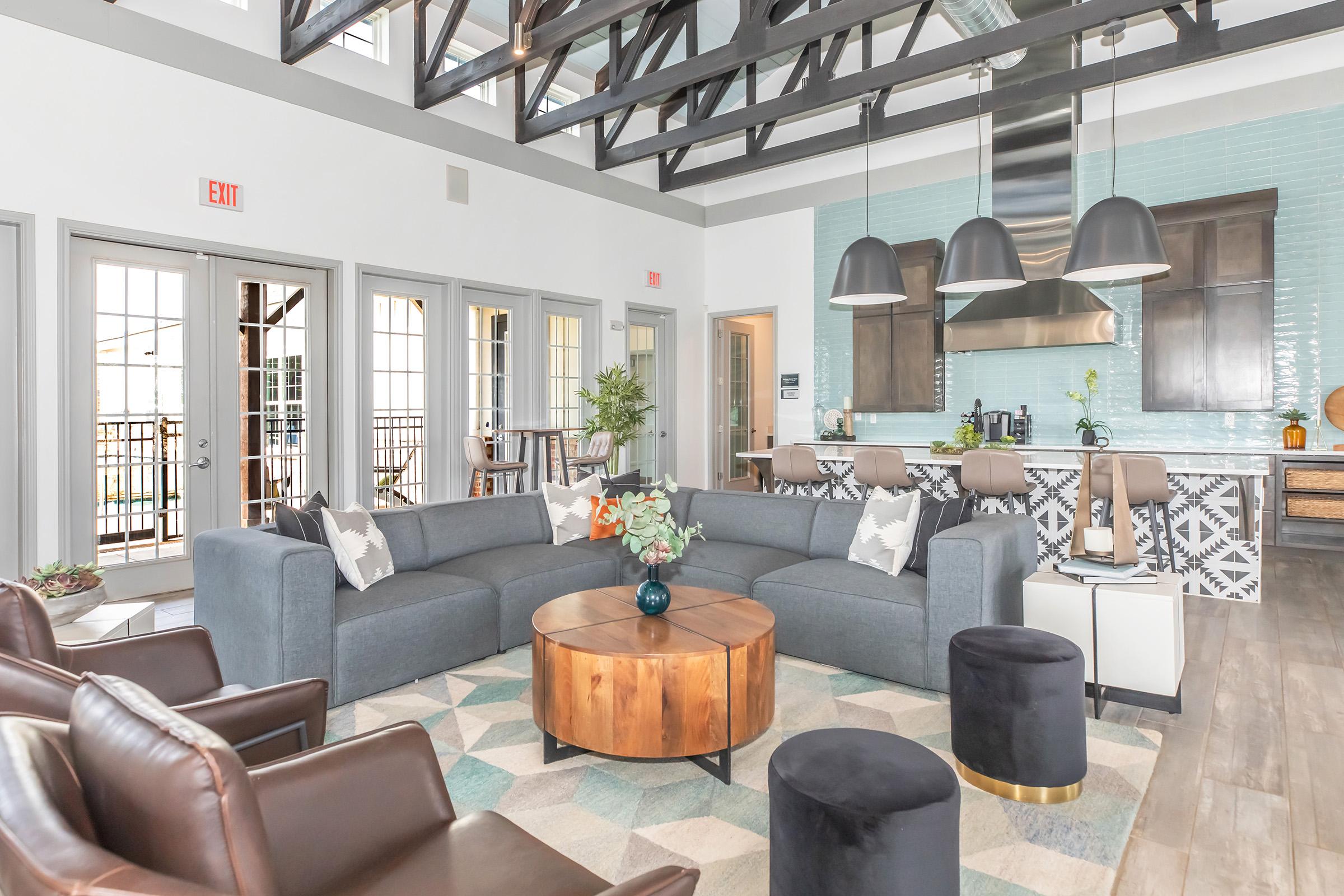 A modern living space featuring high ceilings with exposed beams. The room includes two gray sofas, a round wooden coffee table, and black upholstered stools. A stylish kitchen area with a blue tile backsplash is visible in the background, along with large windows that illuminate the space.
