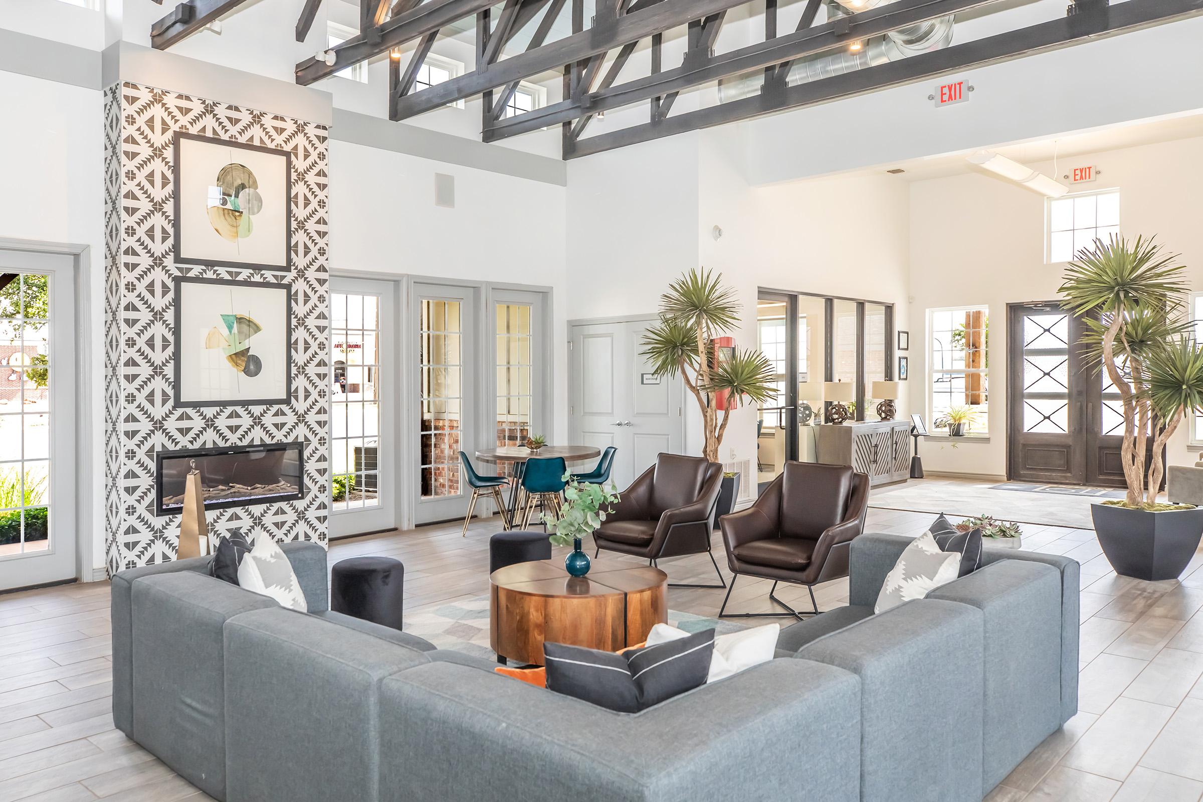 Modern interior of a spacious lounge area featuring a circular wood table surrounded by gray sectional sofas. There are dark leather chairs, potted palm plants, and decorative wall art. Natural light filters in through large windows, and there are wooden beams overhead, creating a stylish and inviting atmosphere.