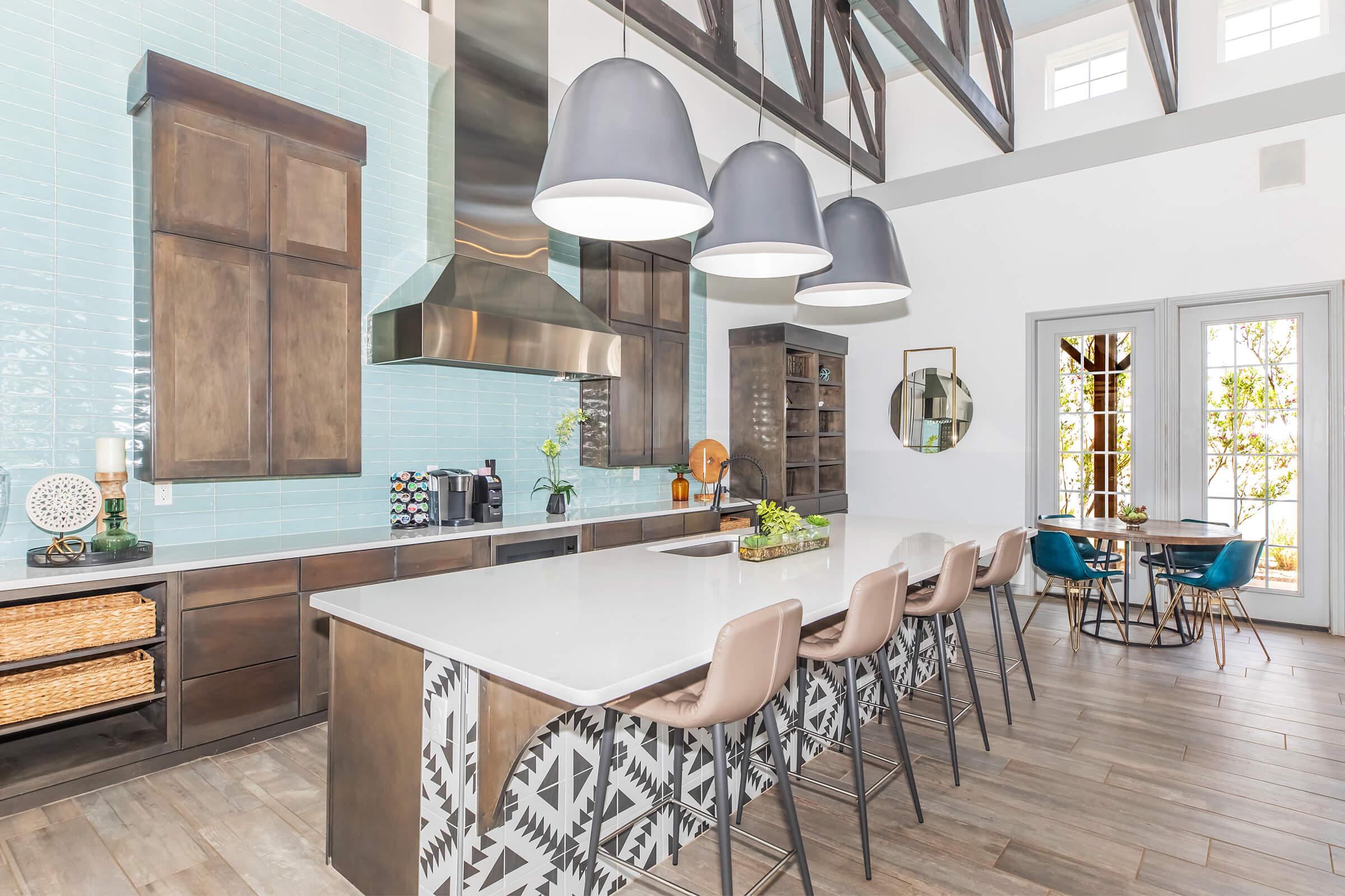 A modern kitchen featuring a large central island with bar stools, sleek cabinets, a stainless steel range hood, and turquoise backsplash tiles. The space has high ceilings with wooden beams, pendant lights, and a dining table with colorful chairs, surrounded by large windows that allow natural light.