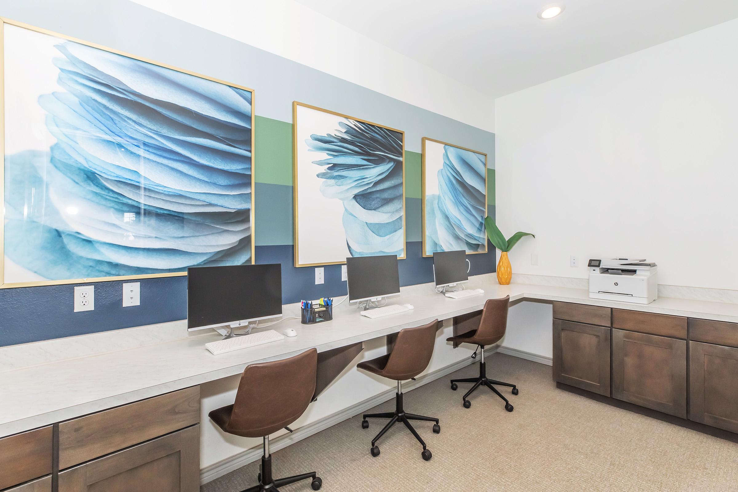 A modern office space featuring three large blue abstract artworks on the wall. There are four computer desks with sleek monitors and rolling chairs. A printer is located on the side, along with a decorative plant and a yellow vase. The overall color scheme includes shades of blue and green.