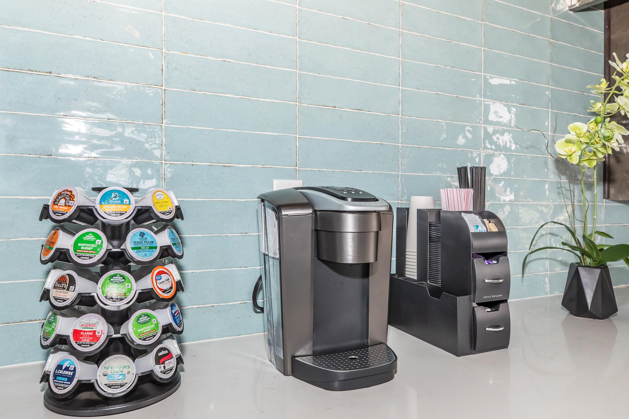 A modern kitchen counter featuring a coffee maker next to a Keurig pod holder with various coffee flavors. To the right, there is a sleek container for cups and straws, and a small potted plant adds a touch of greenery against a blue tiled backsplash.