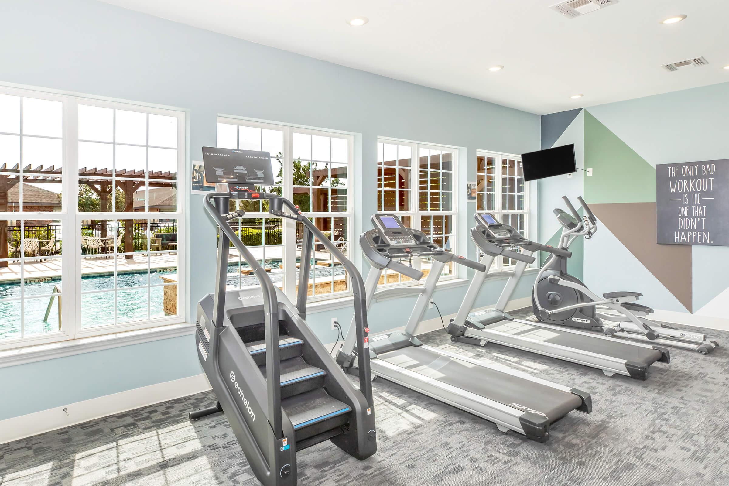 A bright fitness room featuring two elliptical machines and a stair climber, with large windows providing views of an outdoor pool area. The walls are painted in soft colors, and there is a mounted TV for entertainment. The space is designed for exercise and relaxation, bathed in natural light.