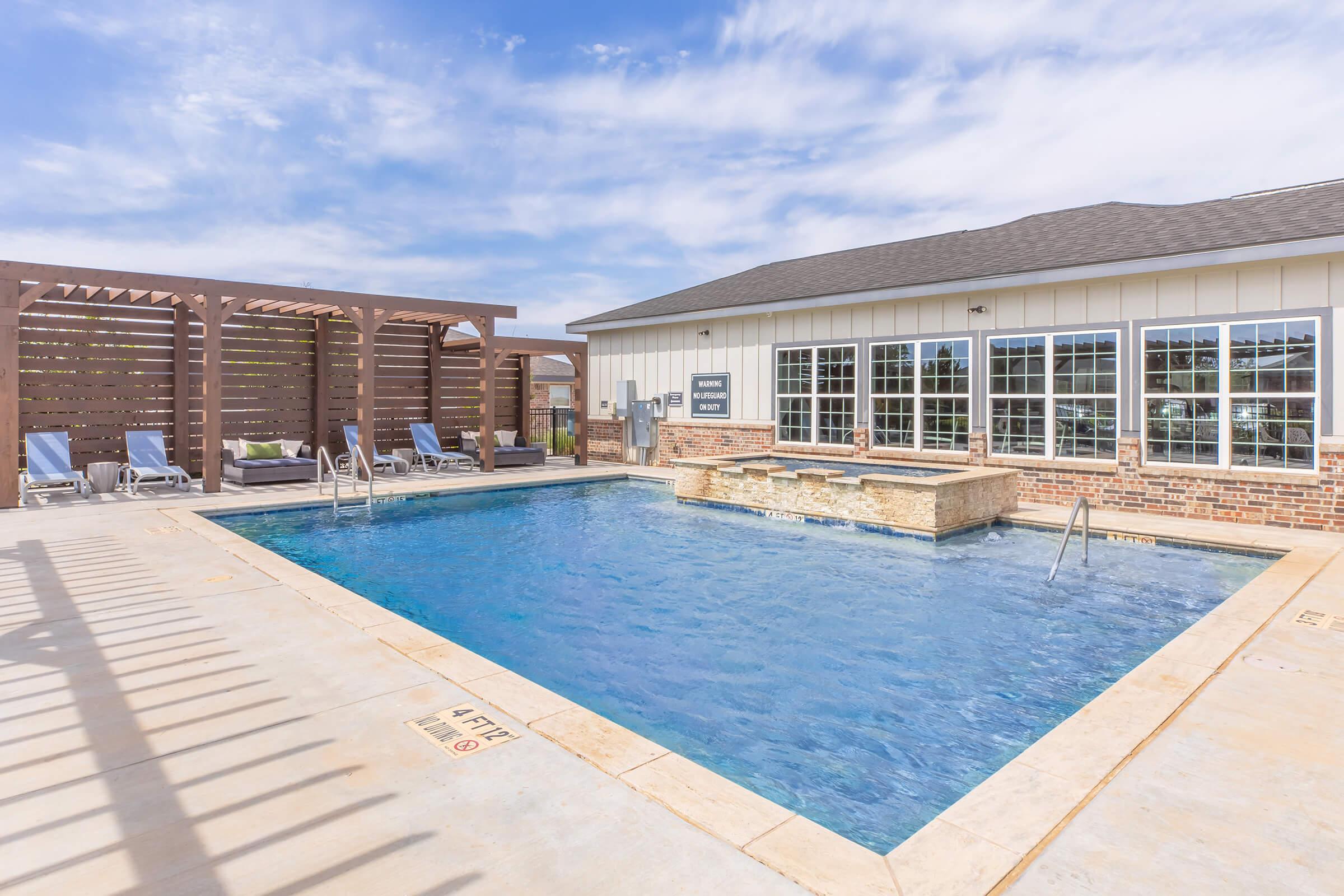A swimming pool featuring an inviting blue surface, surrounded by lounge chairs. A modern building with large windows is nearby, and a wooden pergola provides shaded seating. The area is well-lit under a bright sky with scattered clouds, creating a relaxing atmosphere.
