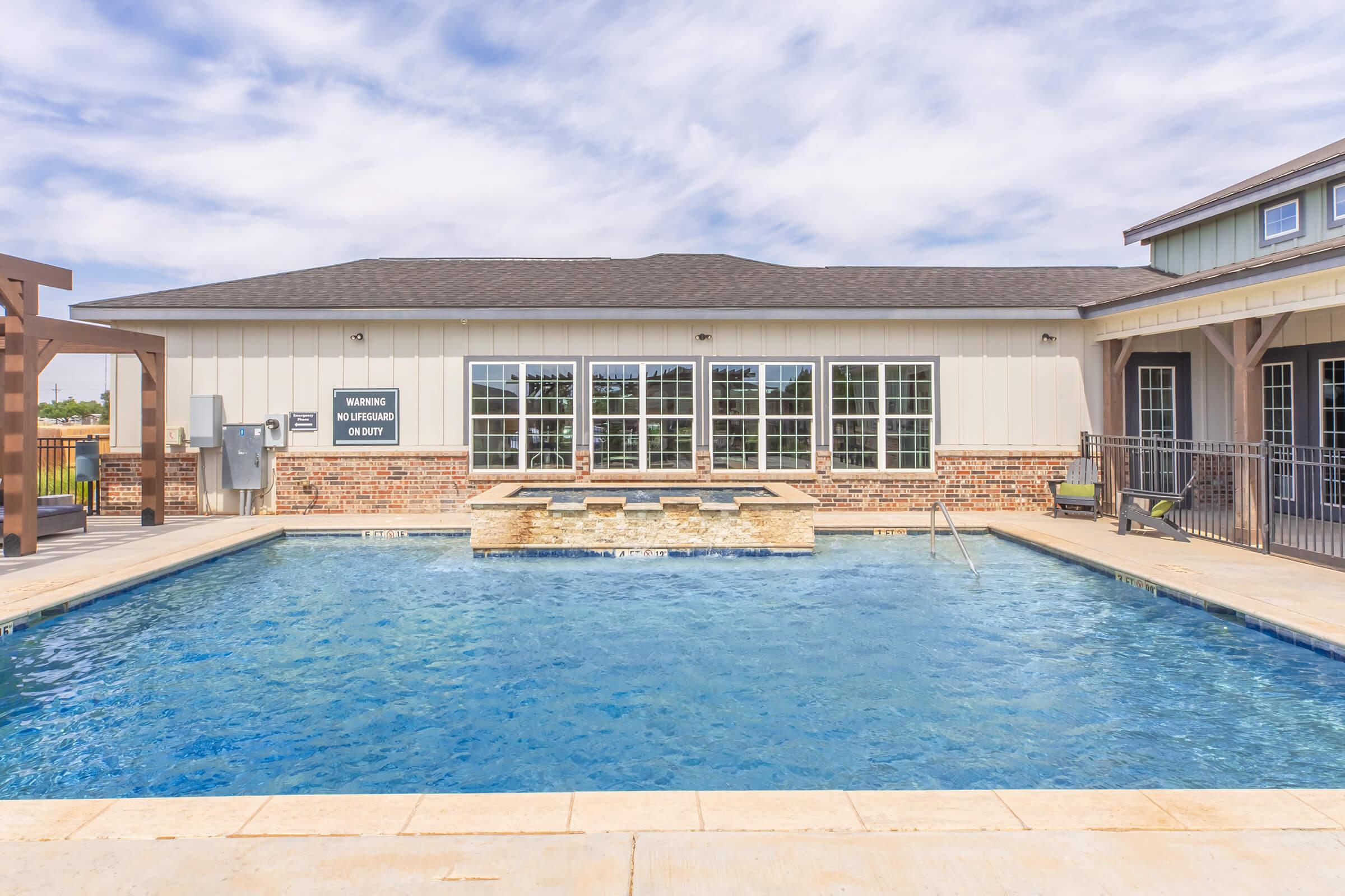 A clear, inviting swimming pool with steps leading into it, located in front of a modern building with large windows. The pool area is surrounded by a stone border and a wooden deck, creating a relaxing outdoor space. The sky is partly cloudy, enhancing the bright atmosphere.