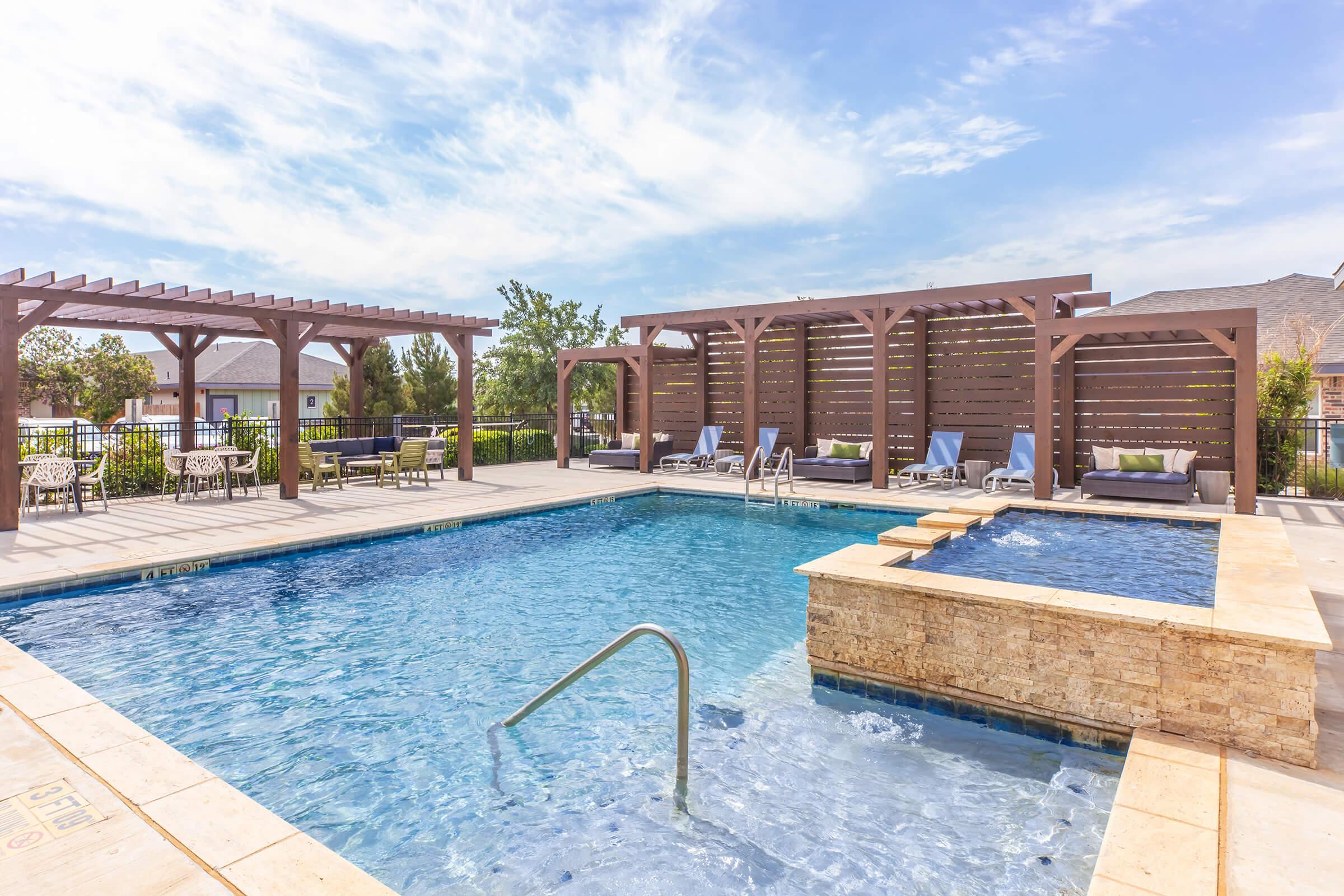 A serene outdoor pool area featuring a clear blue water pool, surrounded by lounge chairs and shaded pergolas. There is a small hot tub adjacent to the pool, set against a backdrop of well-maintained greenery and a few residential buildings under a bright blue sky with scattered clouds.