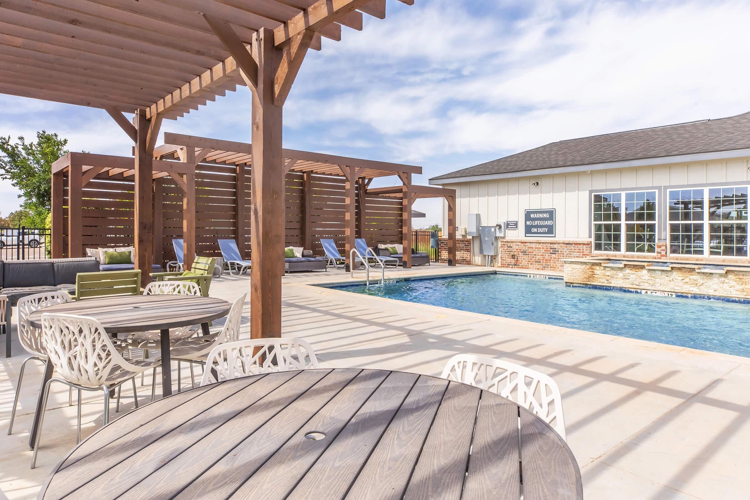 A poolside area featuring a clean, blue swimming pool with lounge chairs and a shaded seating area. In the foreground, there are round wooden tables and white chairs. The background showcases a building with large windows and a pergola, against a clear sky.