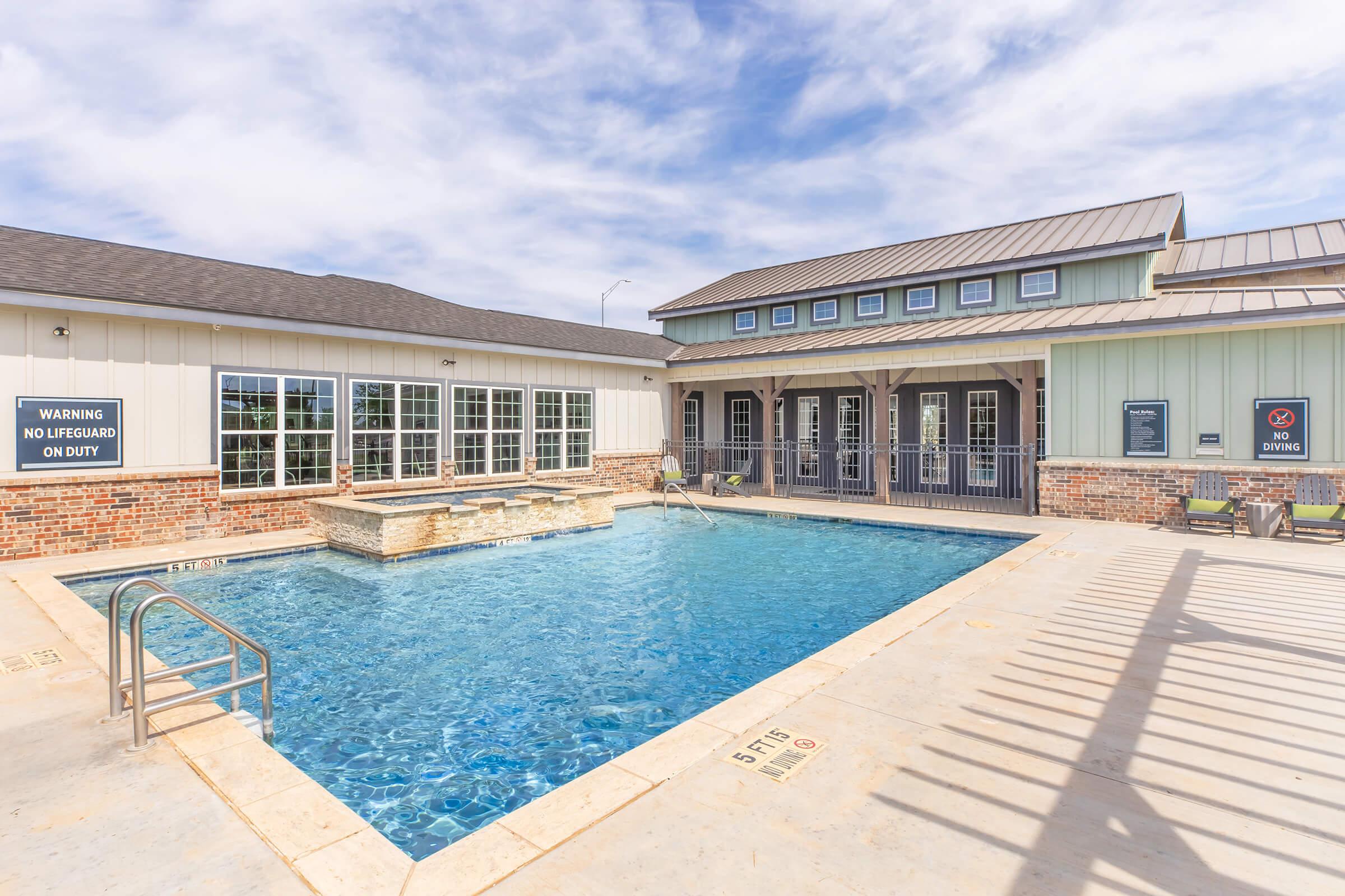 A clear blue swimming pool surrounded by a stone deck, with lounge chairs. In the background, a building features large windows and a sign indicating "No Lifeguard on Duty." The sky is partly cloudy, creating a bright and inviting atmosphere.