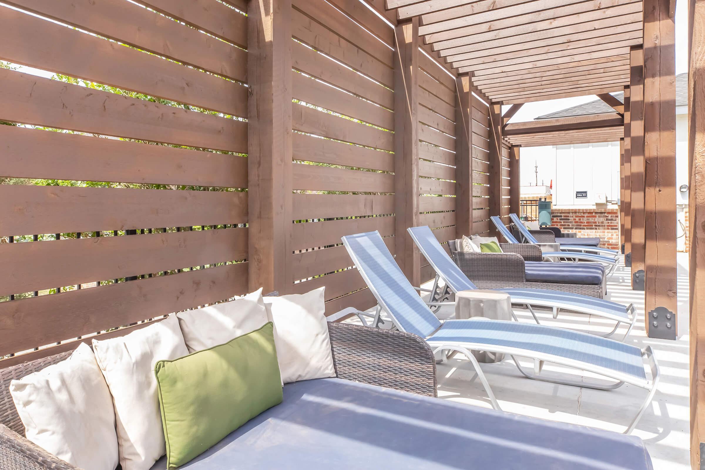 Lounge area with several comfortable chaise lounges under a wooden pergola. The lounges feature cushions in various colors, including green and blue, creating a relaxing outdoor space. Sunlight filters through the slatted wooden structure, enhancing the tranquil atmosphere.