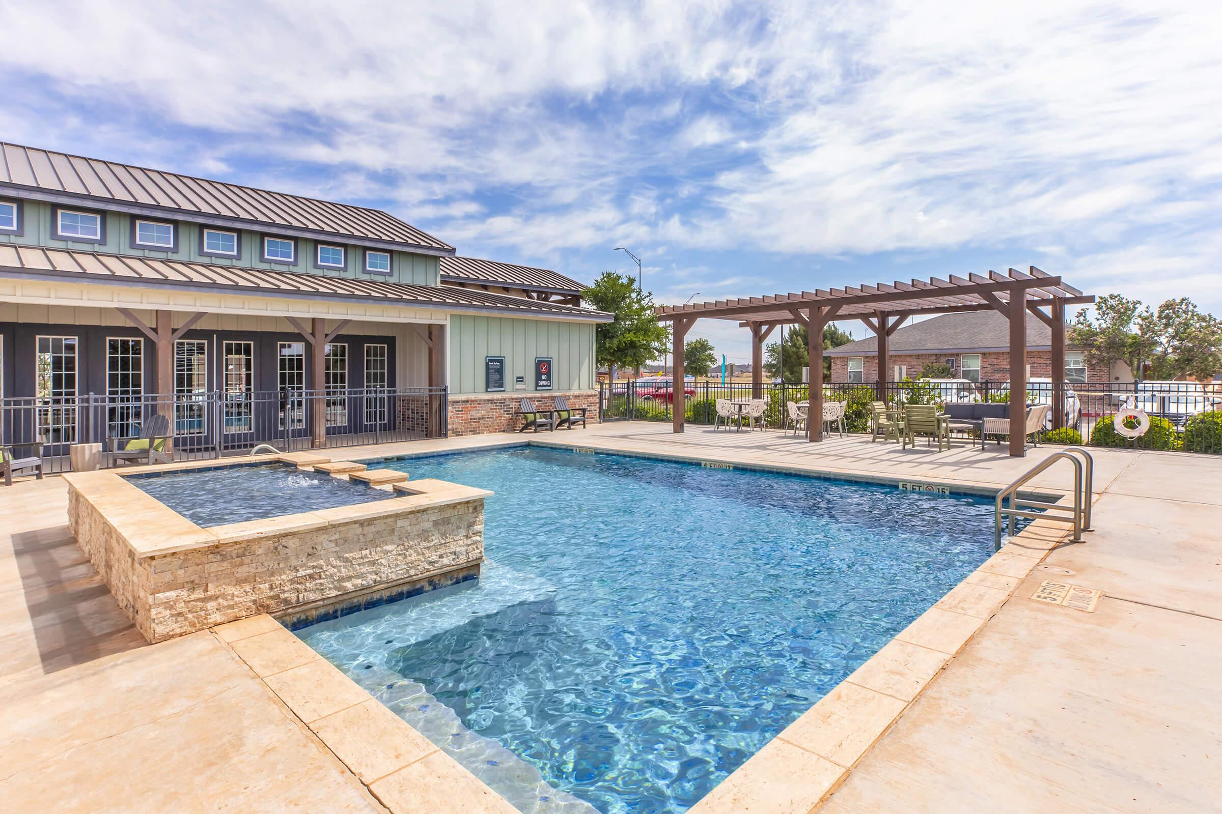 A modern outdoor pool area with clear blue water, surrounded by lounge chairs and tables. A pergola provides shaded seating, and a spa or hot tub is integrated into the pool design. The backdrop features a stylish building with large windows, under a partly cloudy sky.