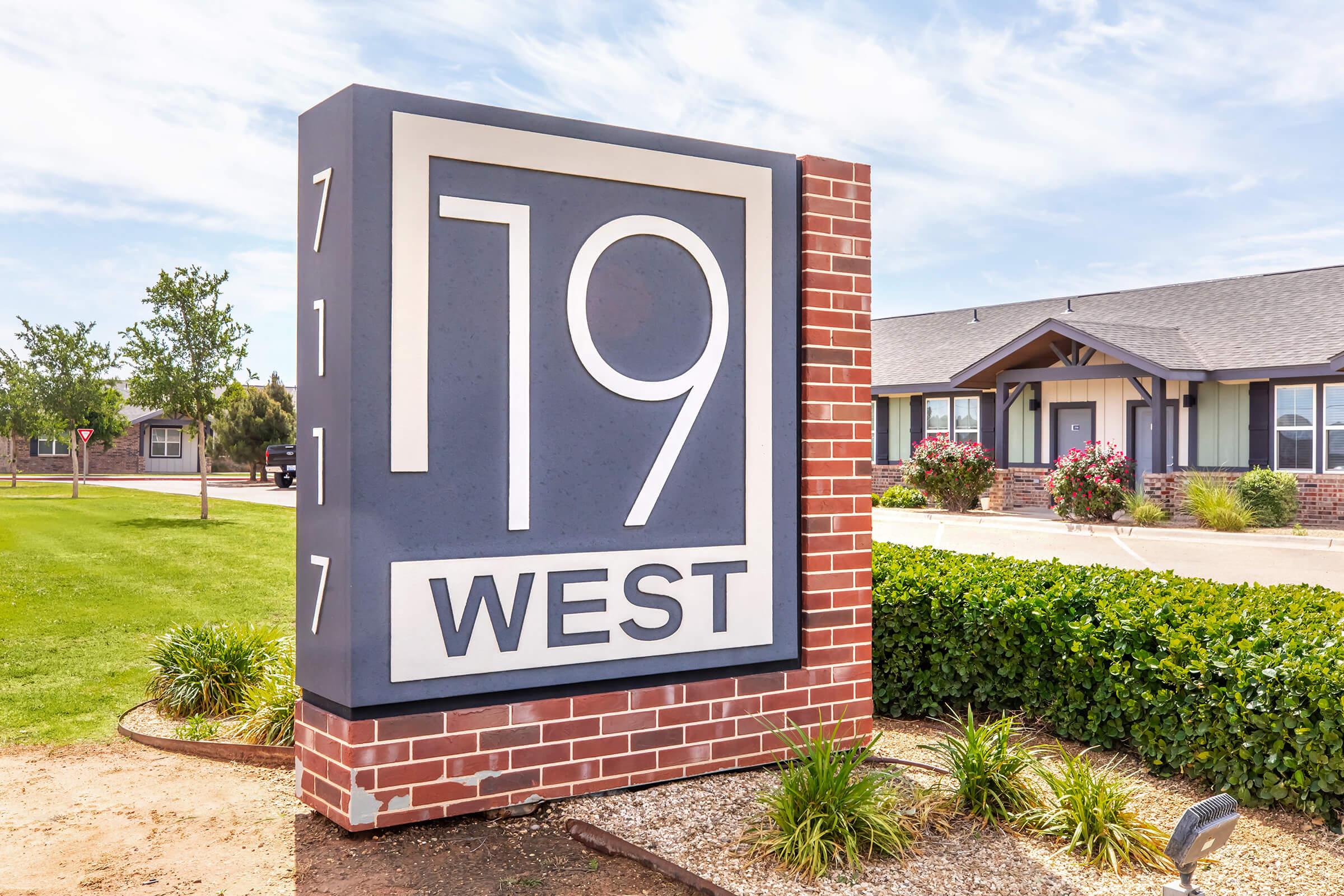 A large, modern sign reading "19 WEST" in bold letters, located at the entrance of a brick building. The sign is surrounded by well-maintained grass and shrubs, with additional greenery in the vicinity, and a clear blue sky in the background. The building features a gray roof and light-colored siding.