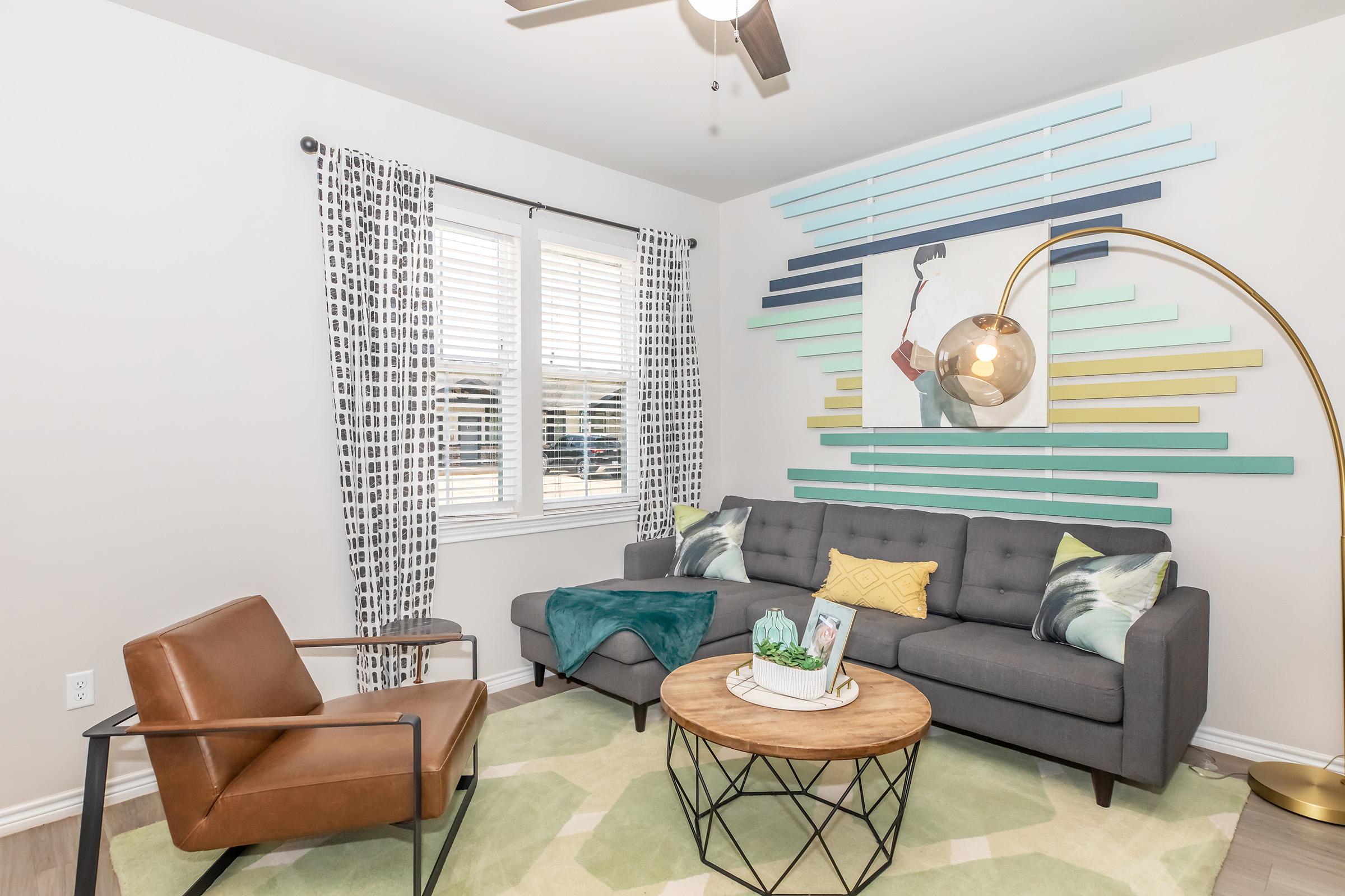 A modern living room featuring a gray sofa with colorful throw pillows, a circular wooden coffee table, and a stylish arc floor lamp. A vibrant wall art piece is mounted above the sofa, complemented by geometric wall decor. Two patterned curtains frame the window, and a cozy green area rug adds warmth to the space.