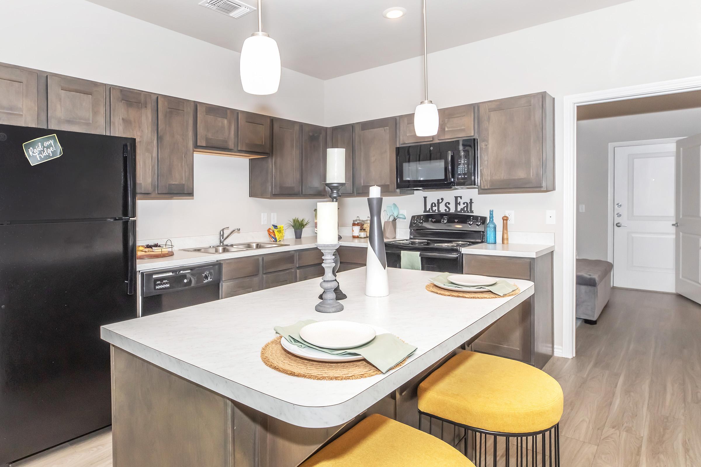 Modern kitchen featuring dark wood cabinets, a black refrigerator, and stainless steel appliances. A white countertop with decorative plates and candles sits on a central island with two yellow bar stools. The space is well-lit with pendant lights, and there's an inviting atmosphere with a door leading to another room.