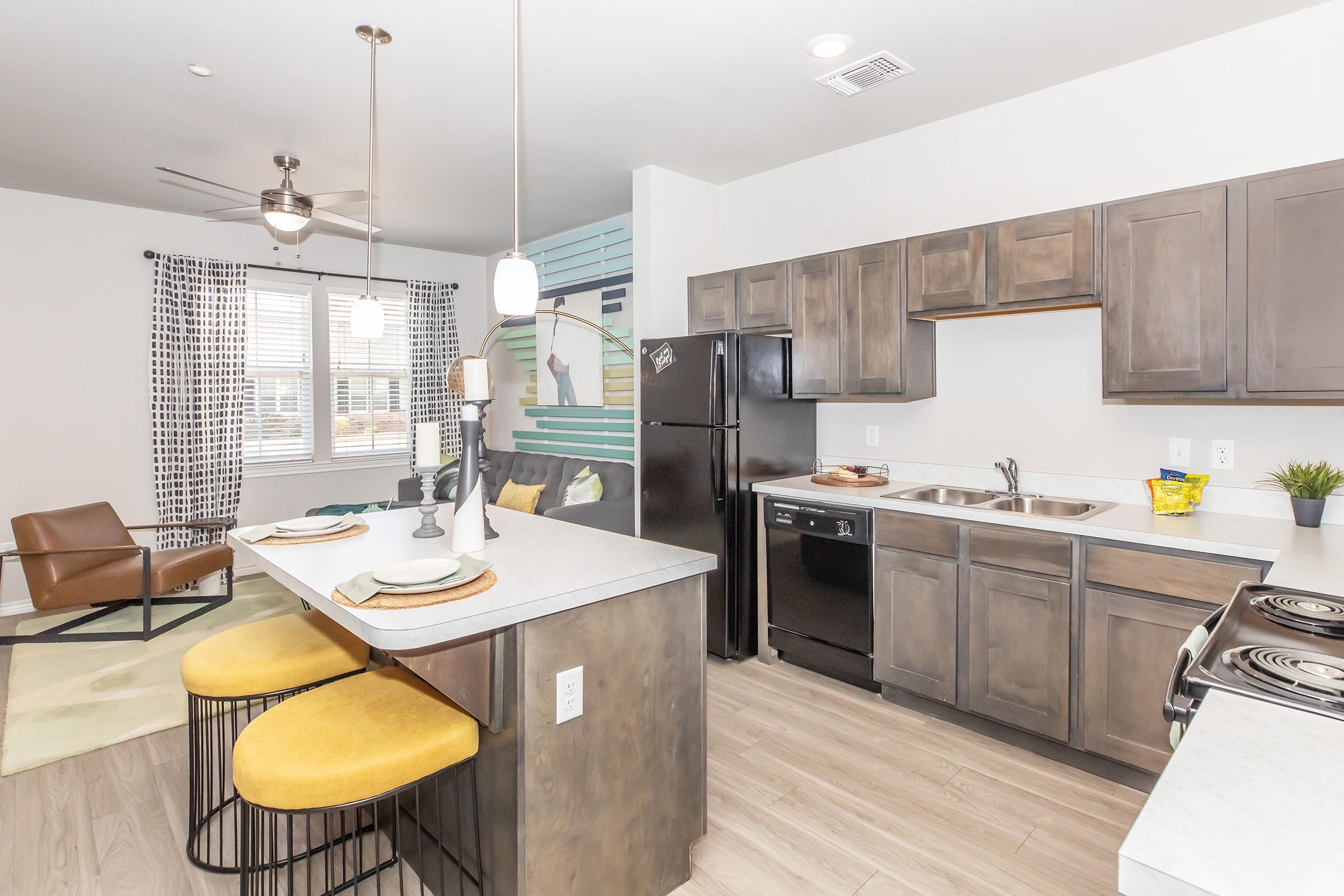 A modern kitchen featuring dark wood cabinetry, stainless steel appliances, and a large island with yellow bar stools. The open-concept design includes a dining area with a light fixture and a comfortable living space with a brown chair and patterned curtains. Natural light fills the room through multiple windows.