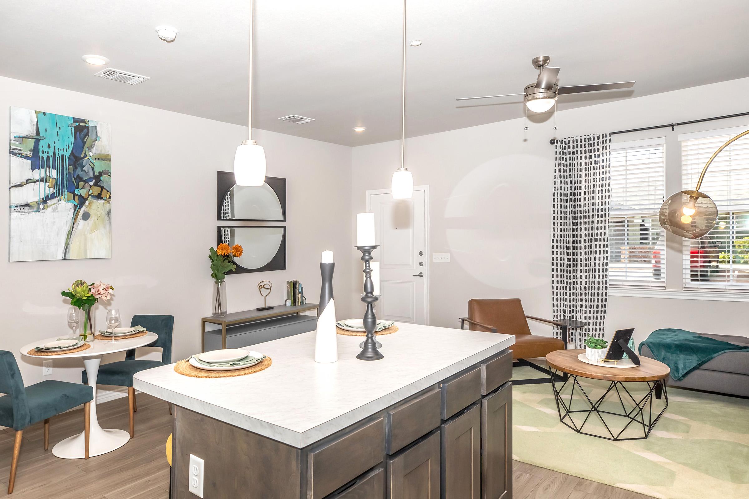 A modern living space featuring a dining area with a small round table and chairs, a comfortable seating area with an armchair and decorative accessories, and stylish lighting. The walls are adorned with artwork, and large windows allow natural light to brighten the room.