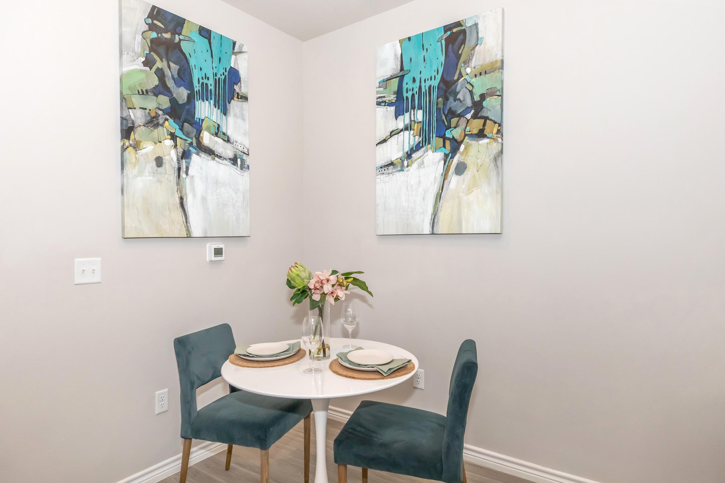 A small dining area featuring a round white table set for two with elegant placemats and glasses. Two plush teal chairs are arranged around the table. Above, there are two large abstract paintings in shades of blue, green, and gold. A vase with fresh pink flowers sits at the center of the table, enhancing the inviting ambiance.