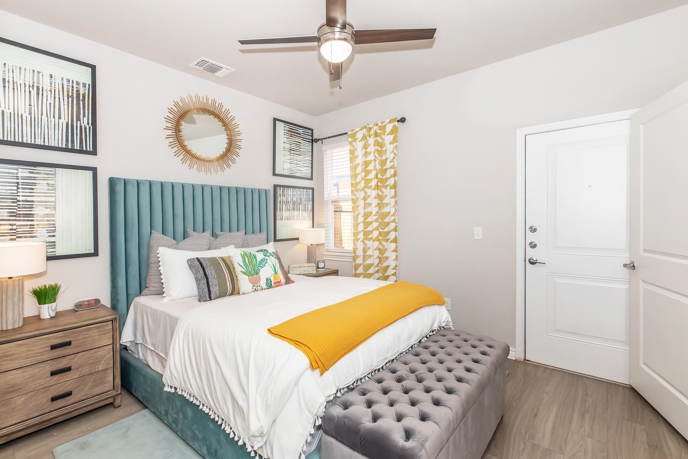 A cozy modern bedroom featuring a plush blue upholstered headboard, white bedding, and a yellow throw blanket. There are decorative pillows, stylish lamps on bedside tables, and a tufted gray ottoman at the foot of the bed. The room has bright curtains and framed artwork on the walls, along with a door leading outside.