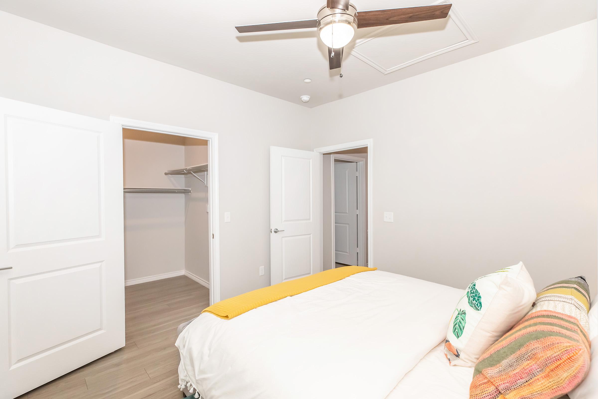 Modern bedroom featuring a bed with colorful pillows and a bright yellow throw blanket. Light gray walls complement the neutral flooring. A ceiling fan provides airflow, and a closet is visible through an open door, adding to the room's functionality. Bright, inviting atmosphere.