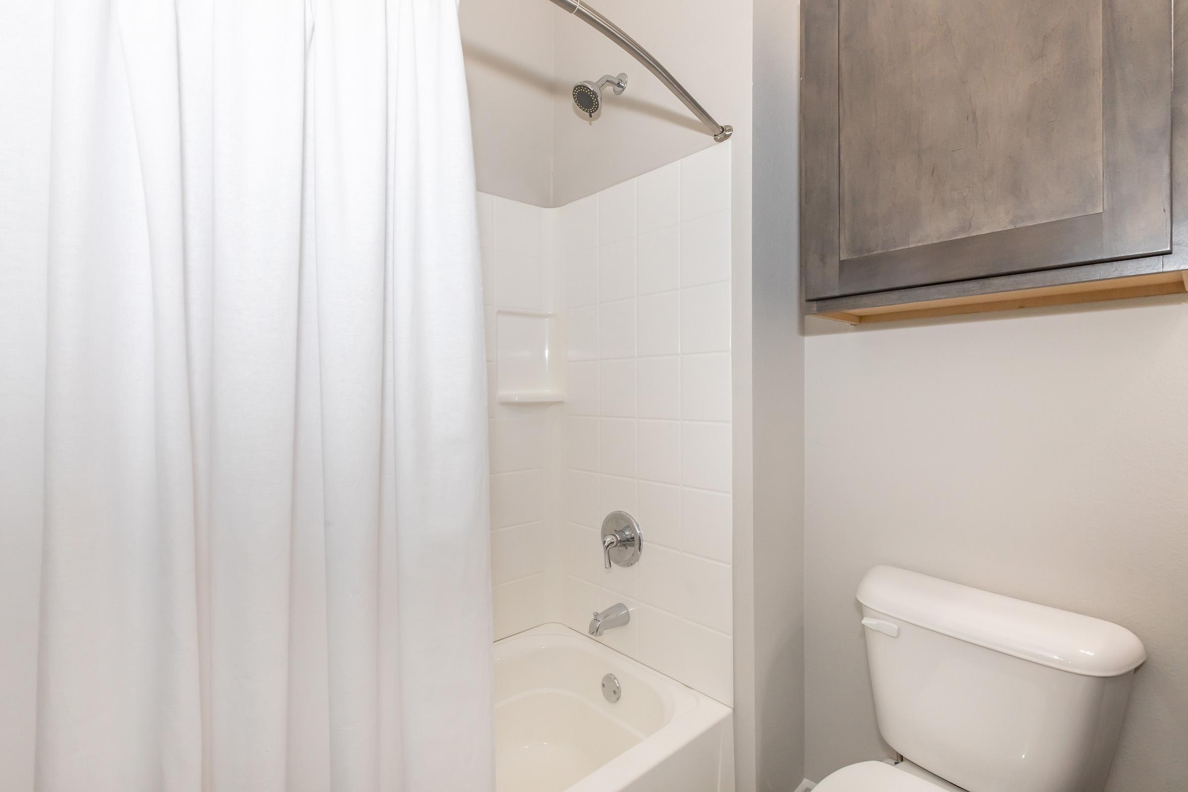 A clean bathroom featuring a shower with a white curtain, a tiled bathtub, a chrome faucet, and a modern toilet. There is a gray cabinet above the bathtub area, with light-colored walls enhancing the spacious look of the room.