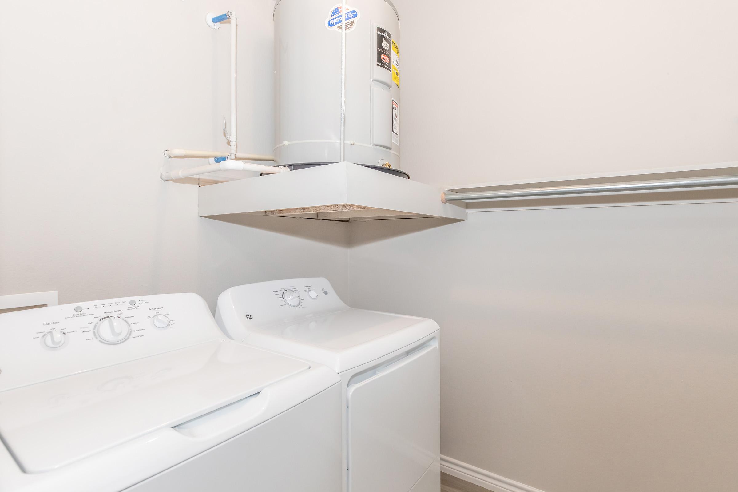 A laundry room featuring a white washing machine and dryer side by side. Above them, a water heater mounted on the wall. The room has light-colored walls and a shelf below the water heater for additional storage or organization.