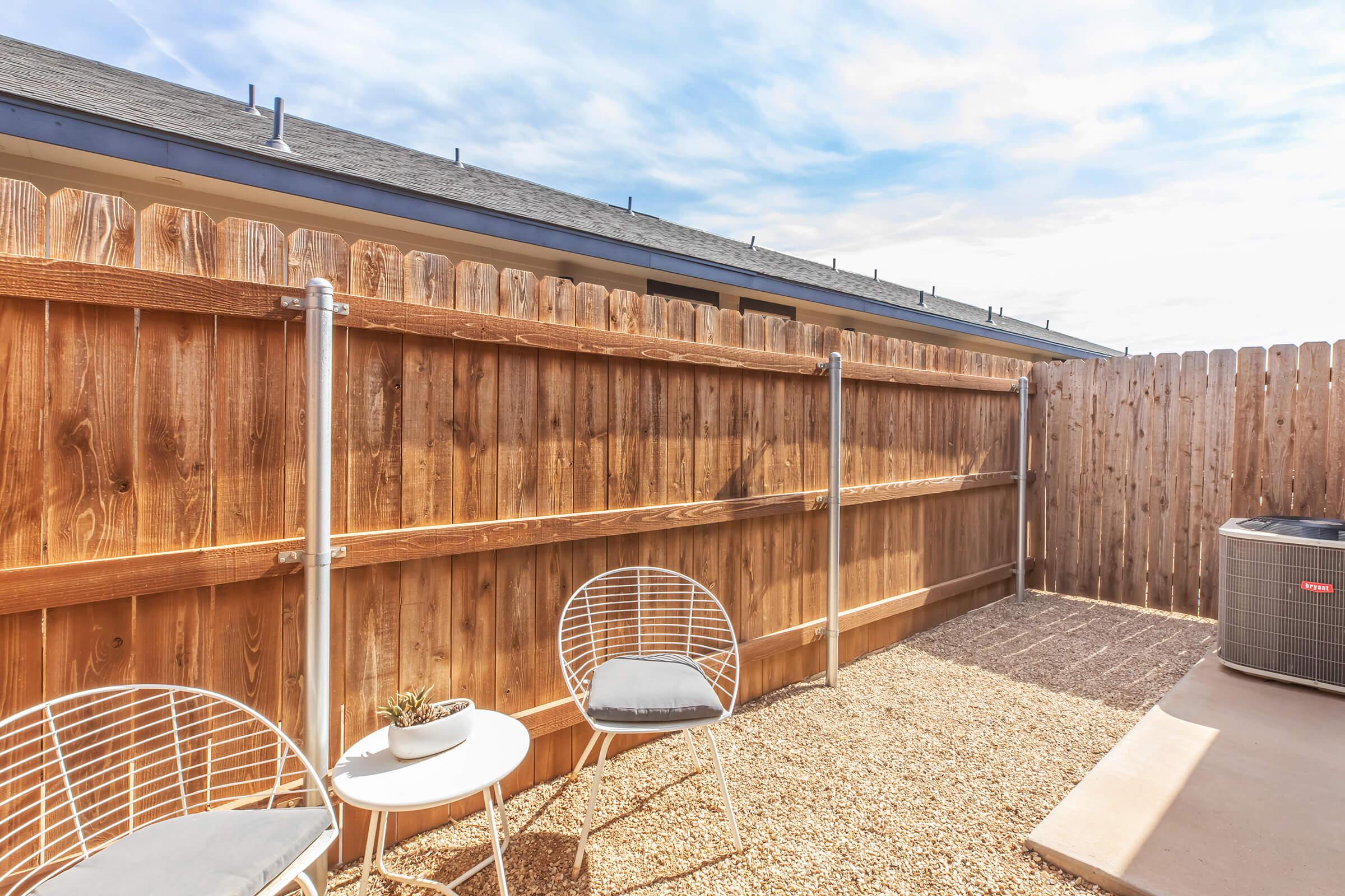 A cozy outdoor space featuring a wooden fence, two white wire chairs with gray cushions, and a small table. A potted plant sits on the table, and an air conditioning unit is visible nearby, surrounded by gravel. The sky is partly cloudy, creating a bright and inviting atmosphere.