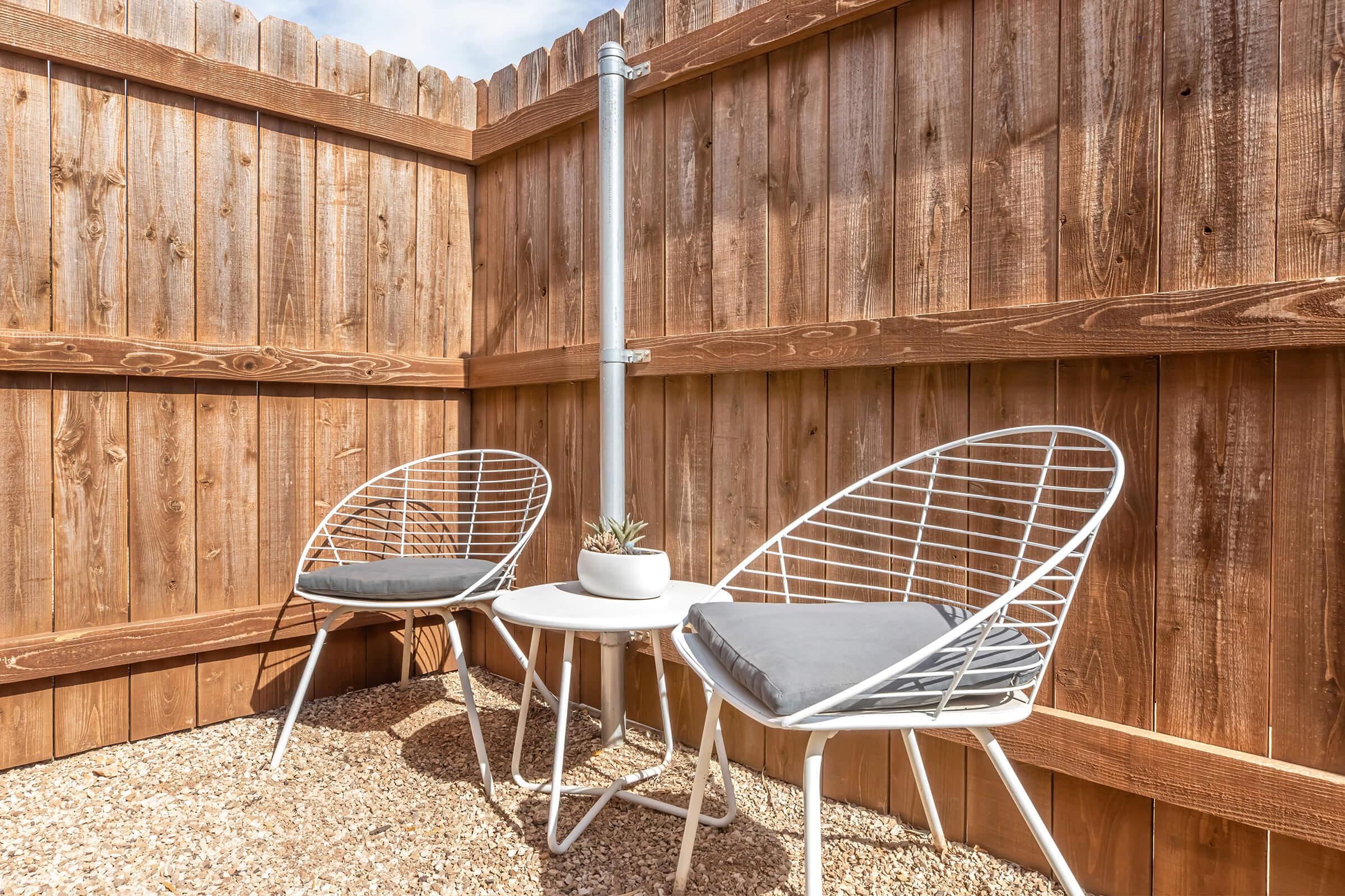 A cozy outdoor corner with two modern white wire chairs and a small round table. The table features a potted plant. The area is enclosed by wooden fencing, and the ground is covered with small pebbles, creating a serene seating space perfect for relaxation.