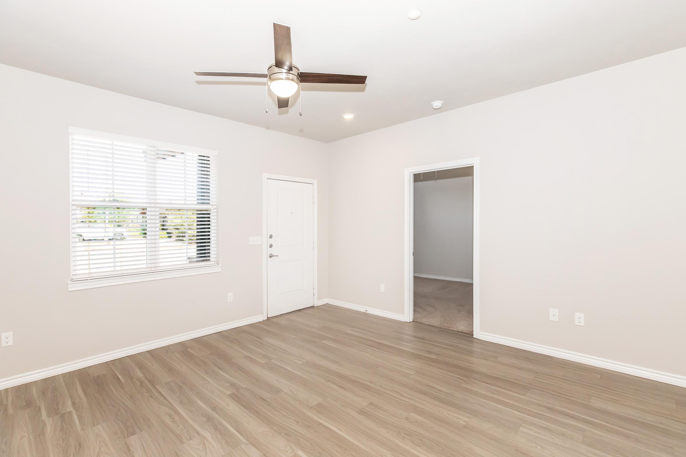 A spacious, empty room with light-colored walls and wood-like flooring. A ceiling fan is visible above. There are white blinds on a window, and a door leads to the outside. A closed door in the background suggests another room, enhancing the sense of openness in the space.