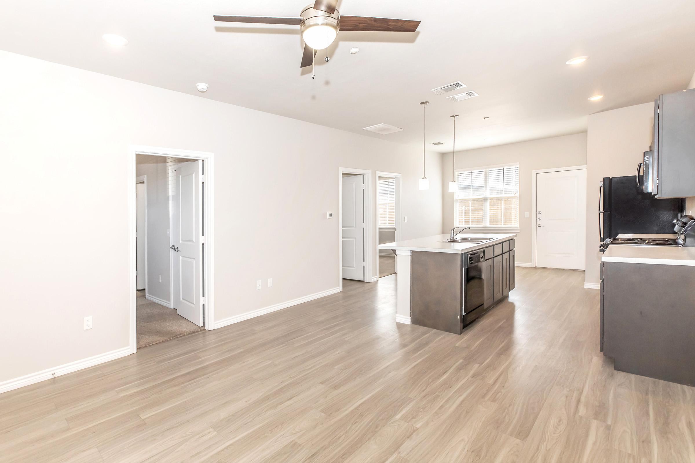 A spacious, modern kitchen with an island and dark cabinetry, featuring a ceiling fan above. Light-colored walls and hardwood floors create a bright atmosphere. Doors lead to other rooms and an entrance, with natural light coming through windows. The area has a clean and inviting ambiance.