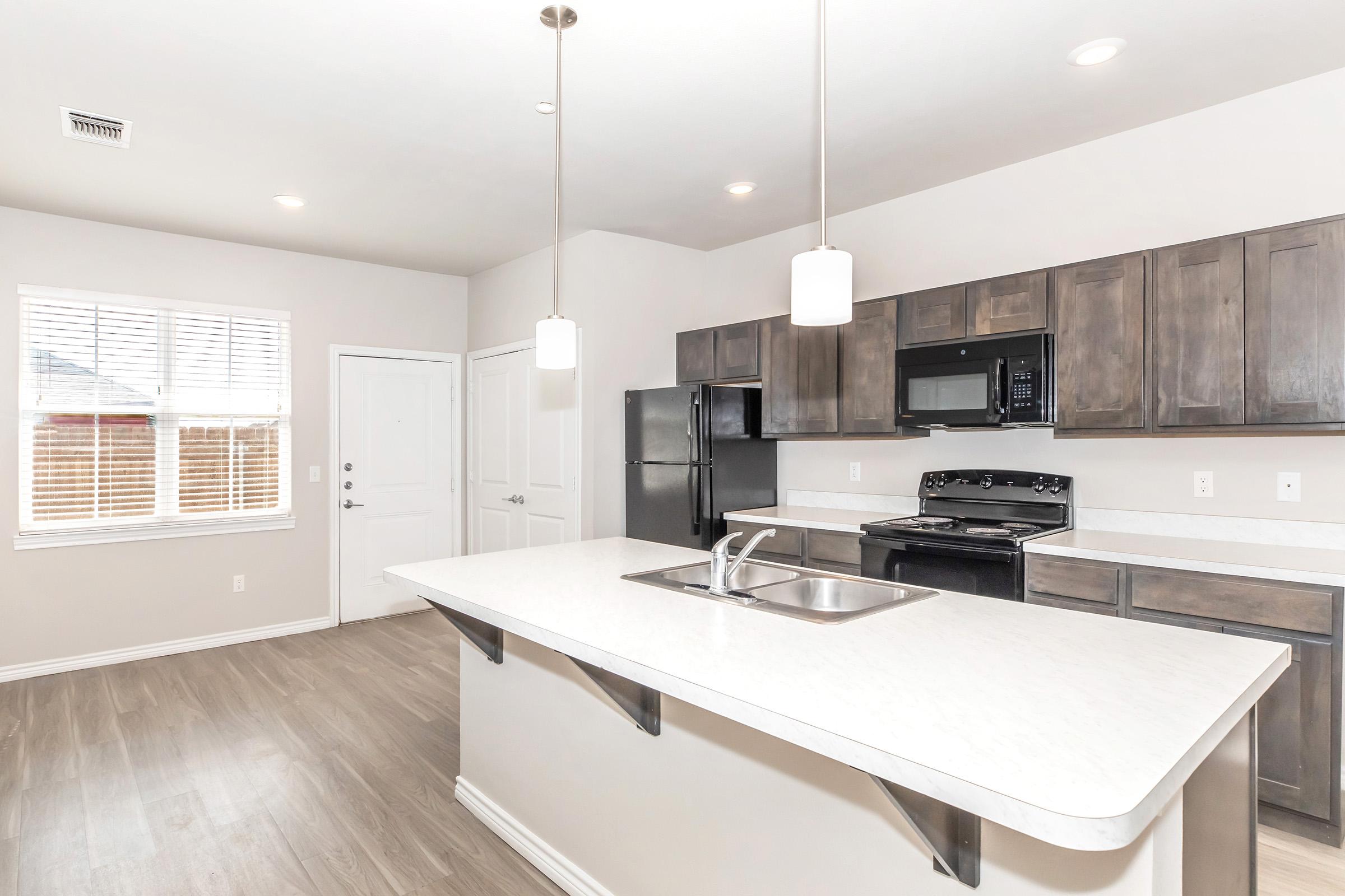 Modern kitchen featuring a stylish countertop island with a sink, dark wooden cabinetry, and stainless steel appliances. The space includes a black refrigerator and microwave, pendant lighting above the island, and a window allowing natural light. A front door is visible, enhancing the room’s openness.