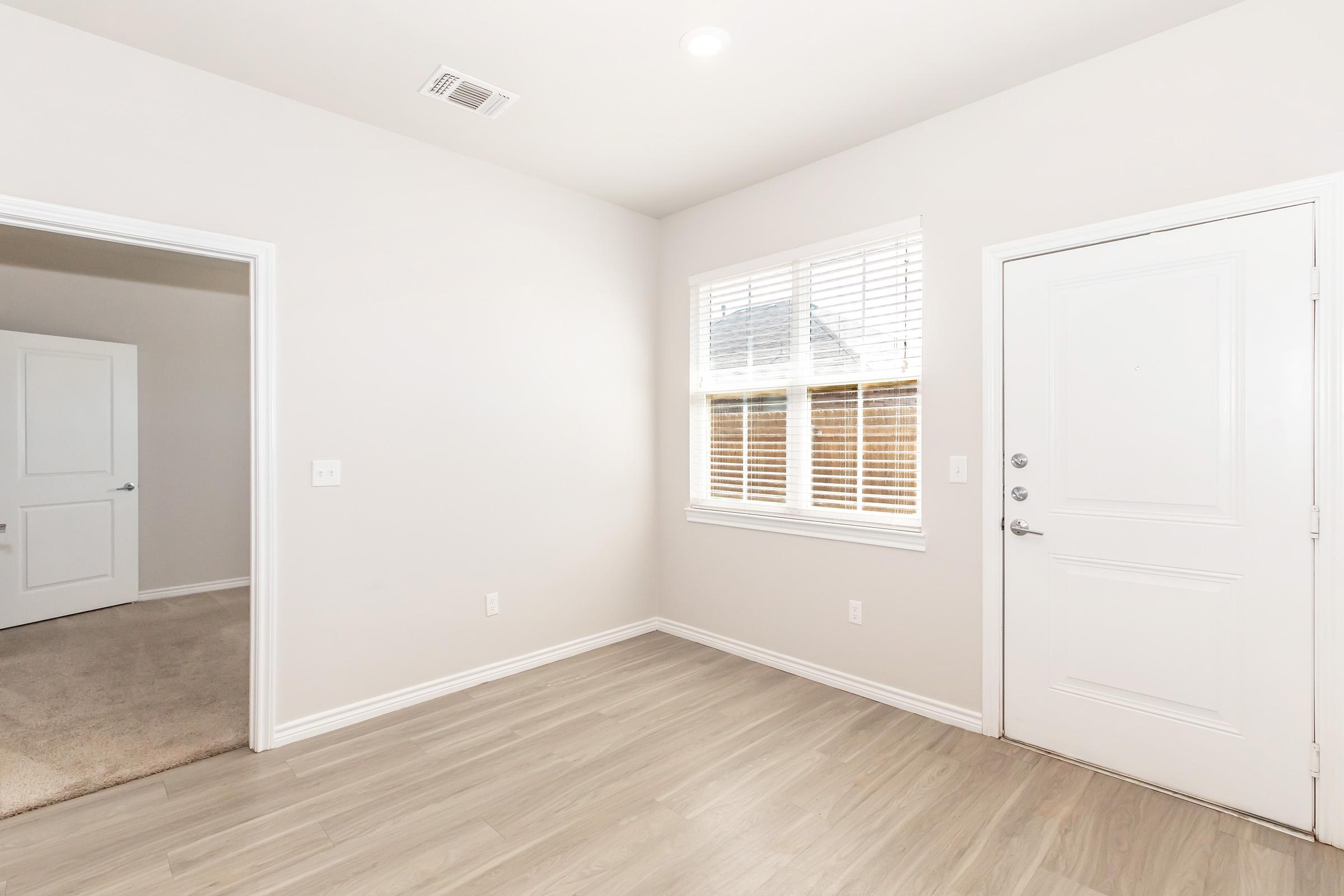 Bright, empty room with light-colored walls and modern trim. A window with closed blinds allows natural light in, while a door leads outside. The room features new flooring and a doorway to another space, creating a clean, open, and inviting atmosphere. Ideal for a living area or small office.