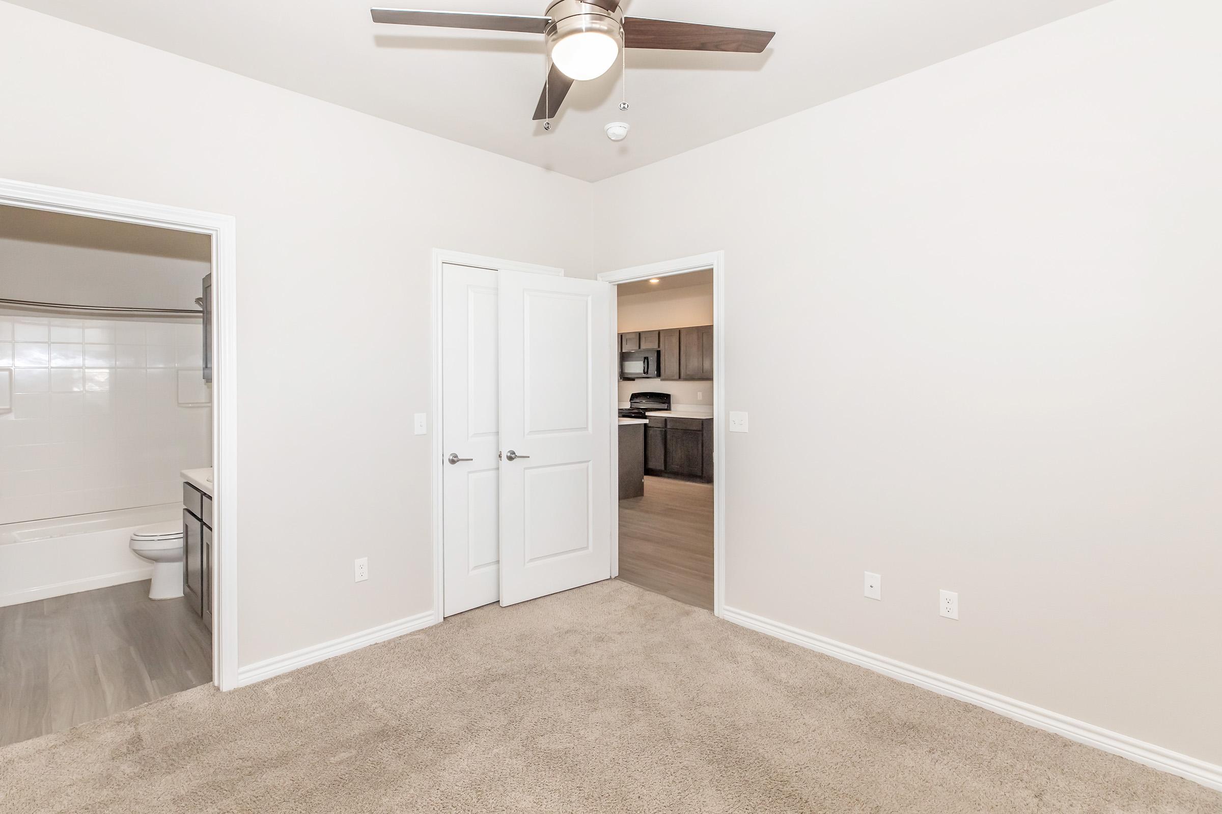 A spacious, light-colored room with beige carpeting, featuring a ceiling fan. Two doors lead to different areas, one possibly to a bathroom, and the other to an adjacent room. The background shows a glimpse of a kitchen with modern cabinetry.