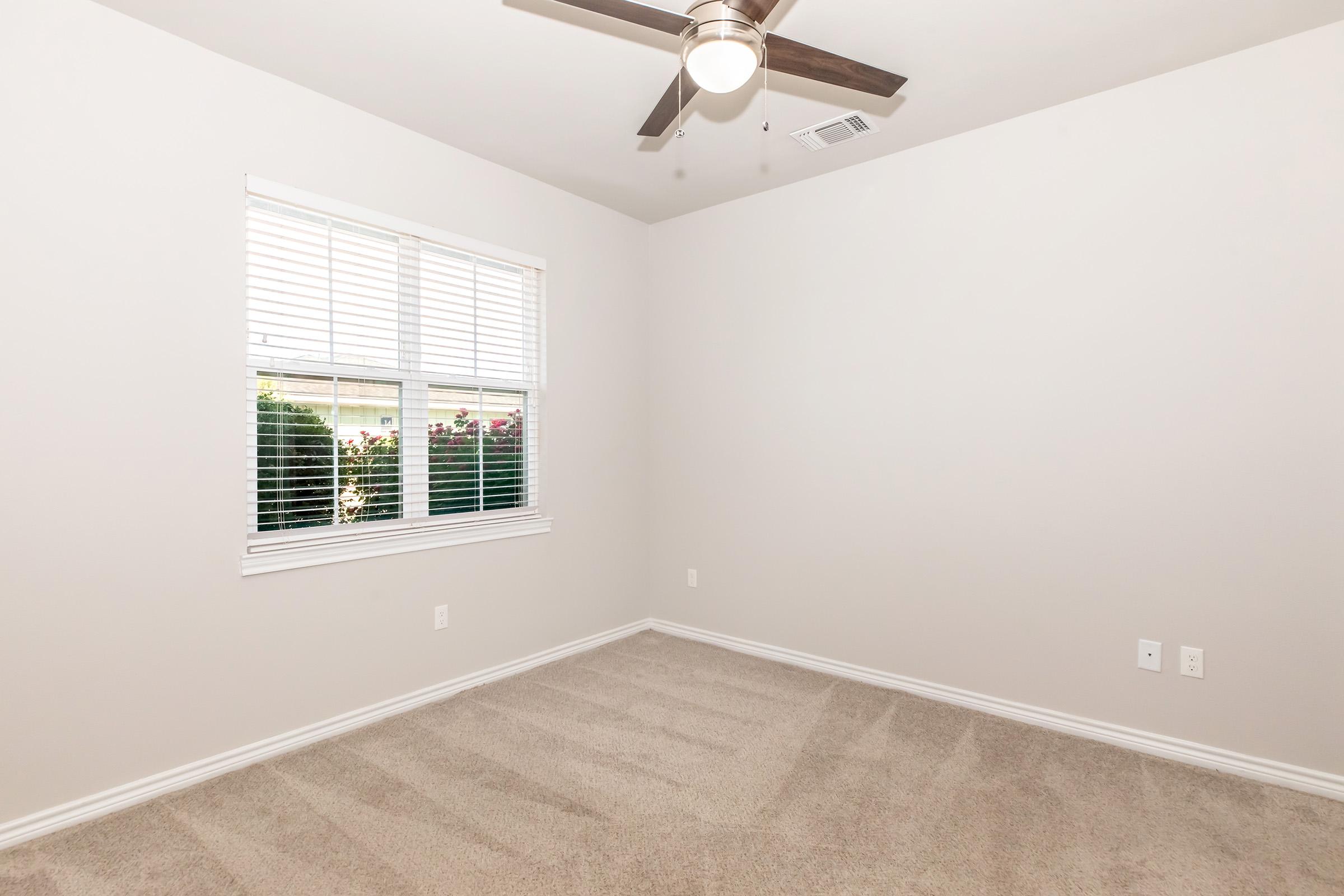 A clean, empty bedroom featuring light beige walls, a ceiling fan, and a window with white blinds. The floor is covered in light-colored carpet, and there are two electrical outlets on the wall. Green plants are visible outside the window, enhancing the natural light in the space.