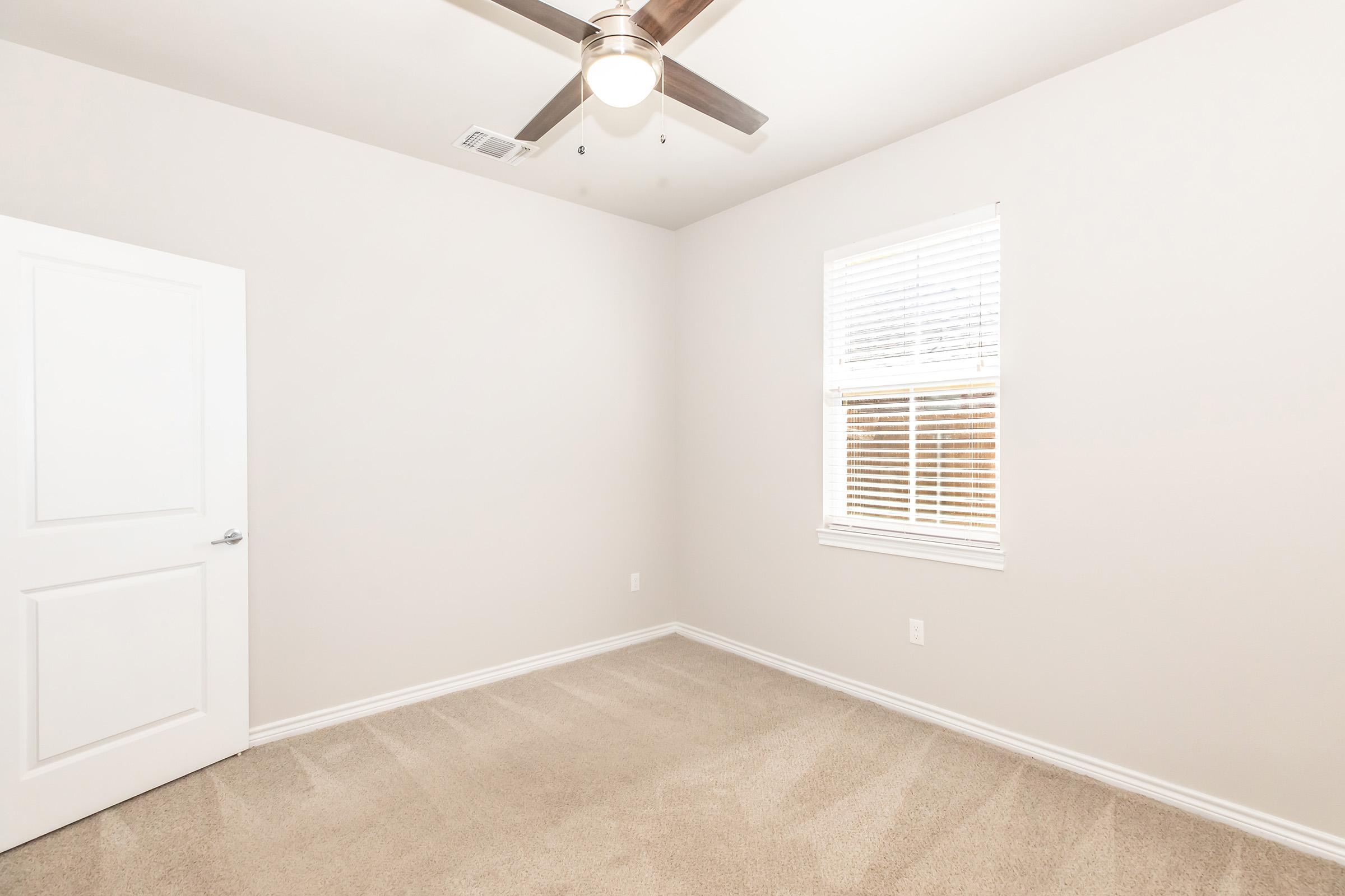 A well-lit, empty room featuring light beige walls, plush carpet, and a ceiling fan. A white door is visible on the left, and a window with blinds is positioned on the right, allowing natural light to enter. The overall atmosphere is clean and uncluttered, suitable for various uses.