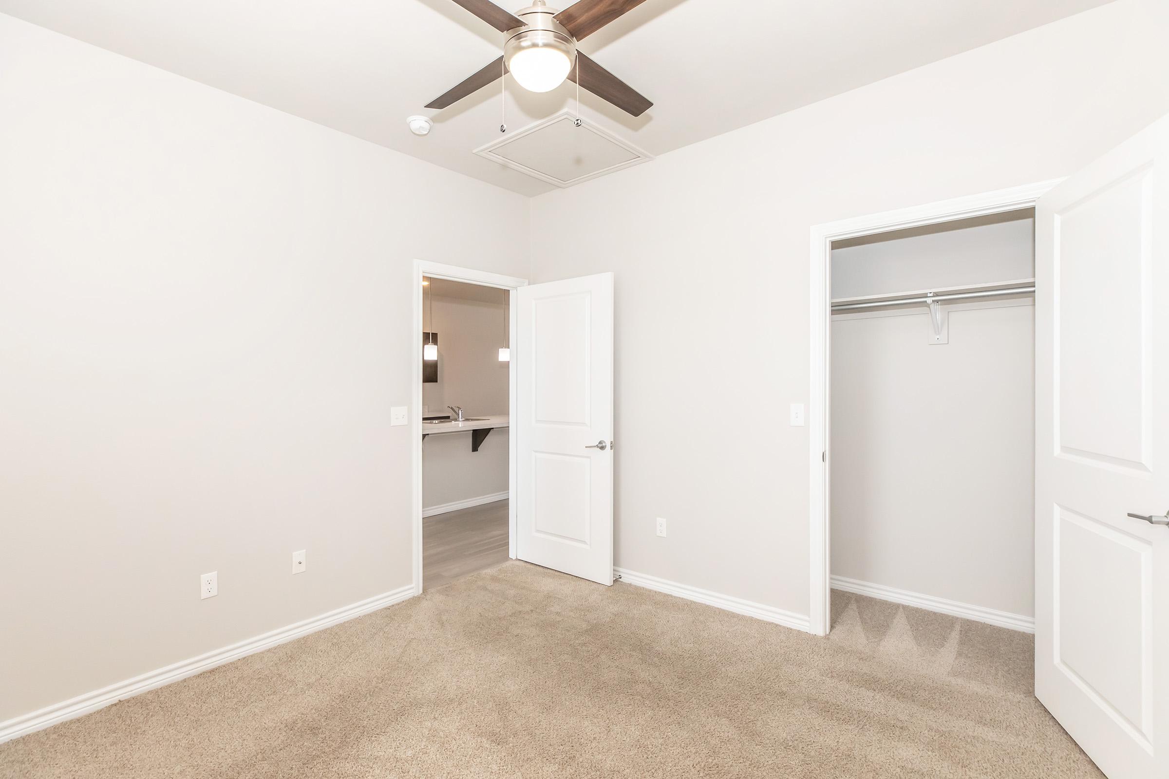 Empty room with light beige carpet, painted white walls, and a ceiling fan. There are two doors: one leading to a closet and the other possibly to an adjoining room. The room is well-lit and spacious, with natural light coming from an unseen window.