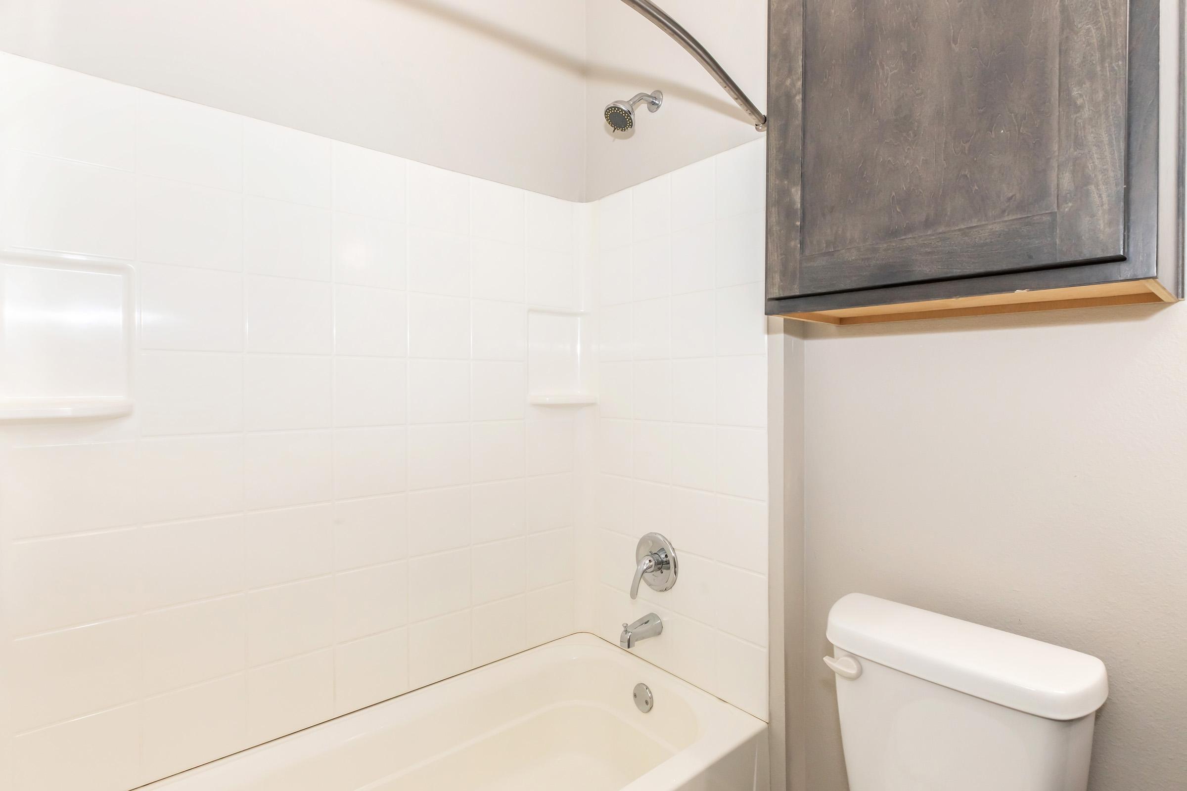 A clean bathroom featuring a white tiled shower area with a bathtub, a chrome showerhead, a wall-mounted storage cabinet, and a white toilet. The walls are a neutral shade, complementing the fixtures and enhancing the overall brightness of the space.