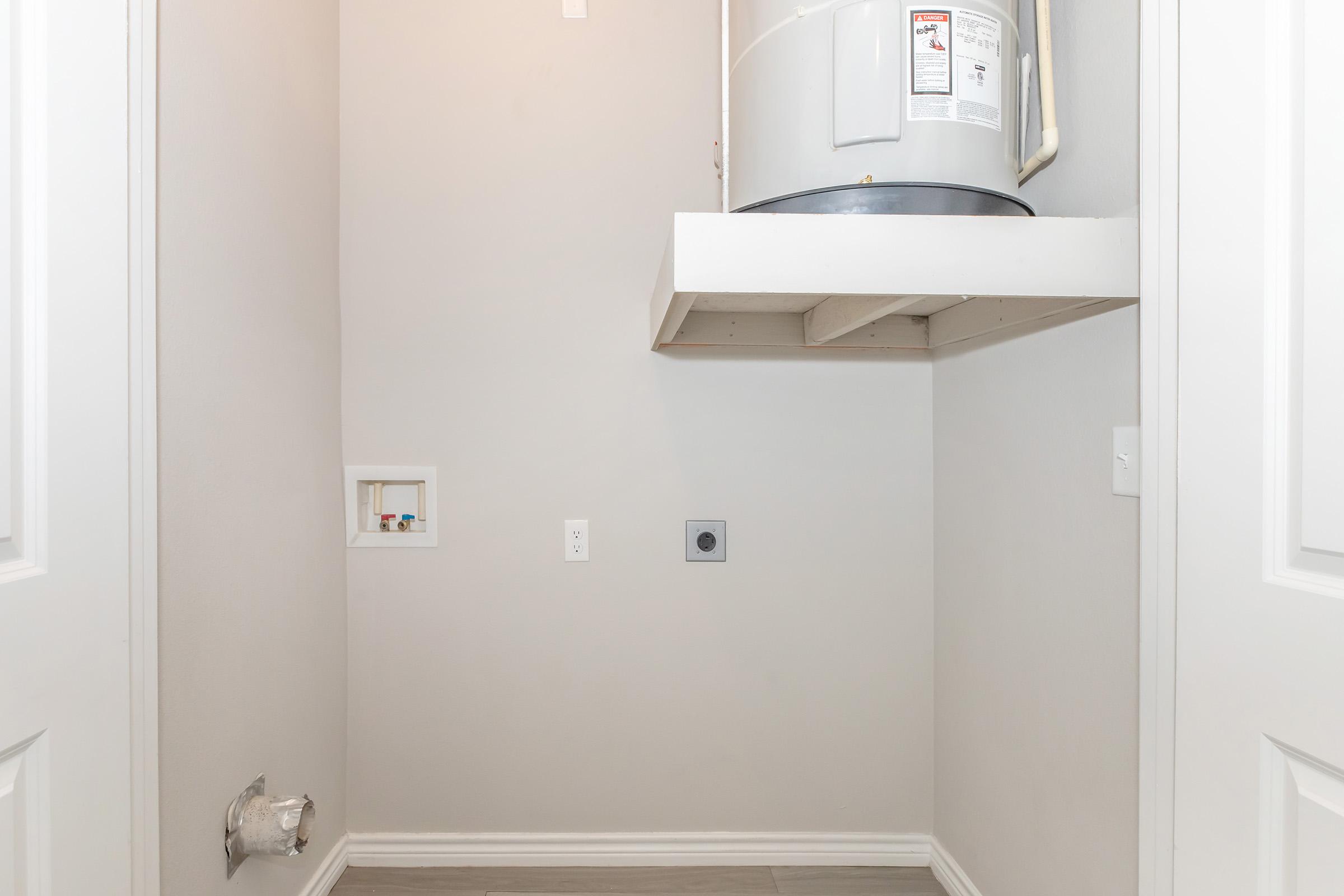 A sparse laundry room with light gray walls, featuring a water heater mounted on a shelf. There are utility connections visible on the wall, an outlet, and a vent at the floor level. The space is well-lit and has two closed doors, suggesting it’s a small, functional area for laundry appliances.