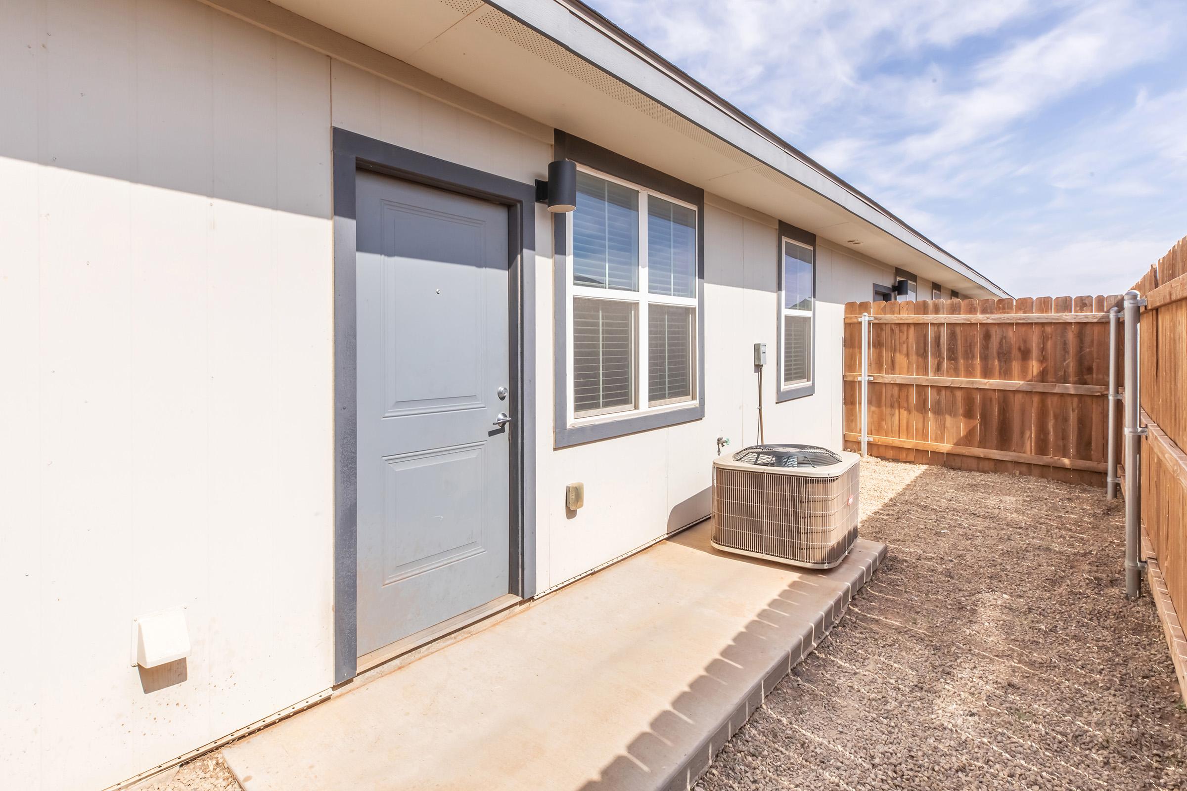 Side view of a single-story building featuring a gray door, windows, and an air conditioning unit. The area is enclosed by a wooden fence with a gravel surface. The sky is partly cloudy, adding to the exterior ambiance of the property.