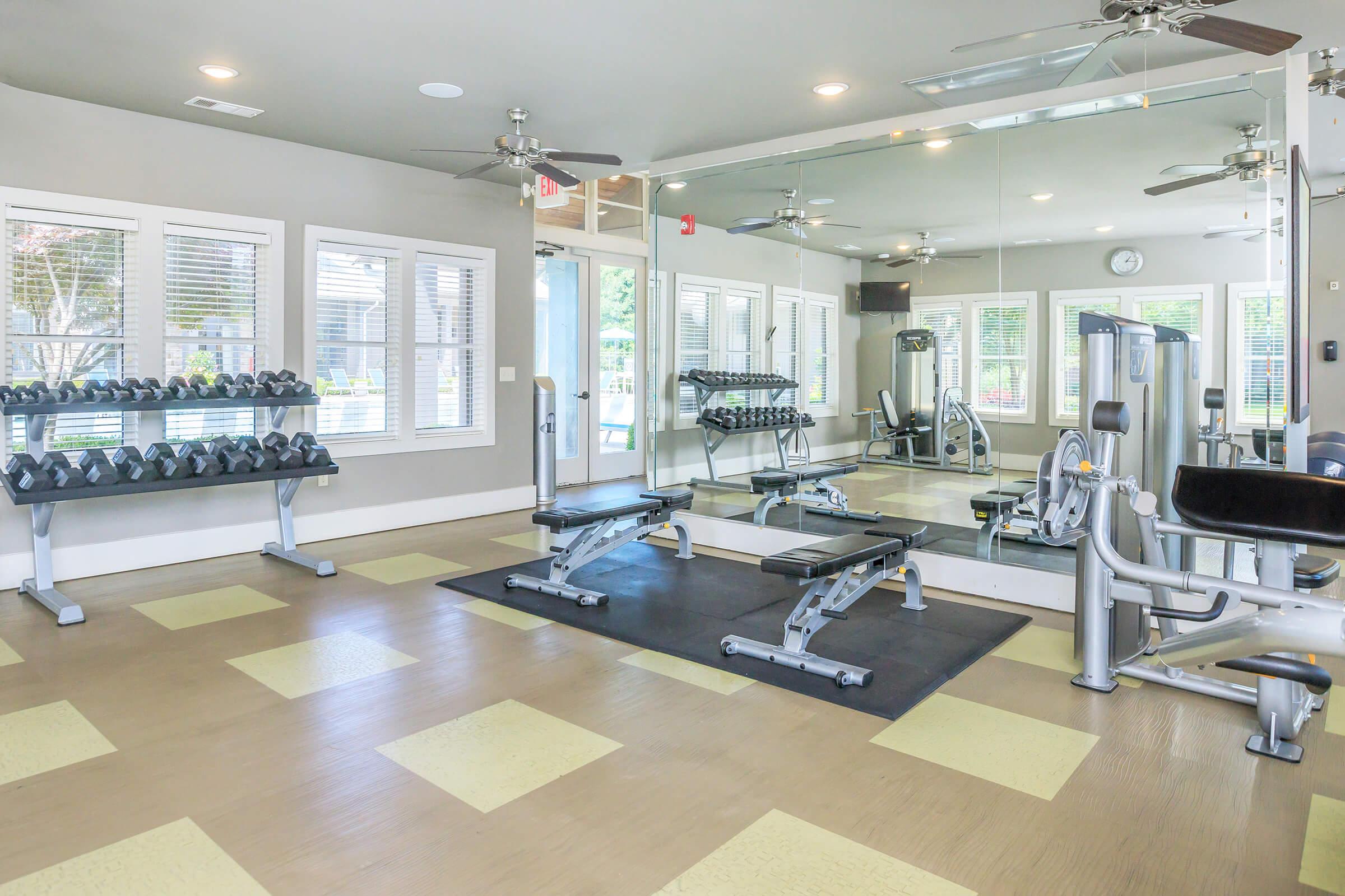 A bright and modern gym interior featuring a weight rack with dumbbells, a multi-use weight bench, and various workout machines. Large mirrors reflect the workout area, which has several windows allowing natural light to enter. The floor has a combination of yellow and beige tiles, creating a welcoming atmosphere for exercise.