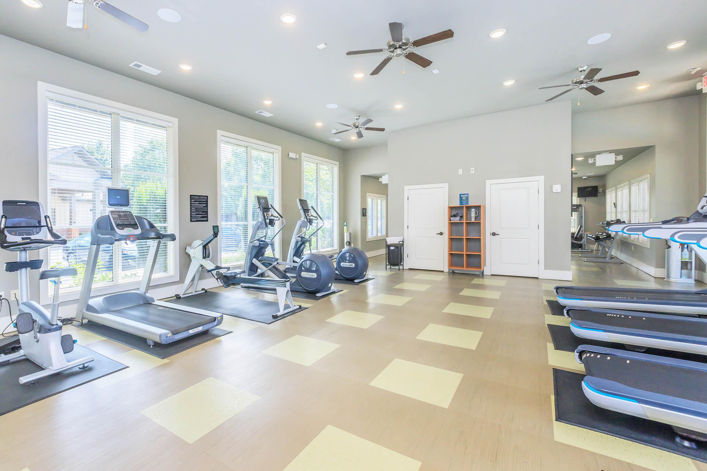 A modern gym interior featuring various exercise equipment, including treadmills, elliptical machines, and stationary bikes. The space is well-lit with multiple ceiling fans and large windows allowing natural light. There are also benches and storage cubbies along the wall, creating a clean and organized workout environment.