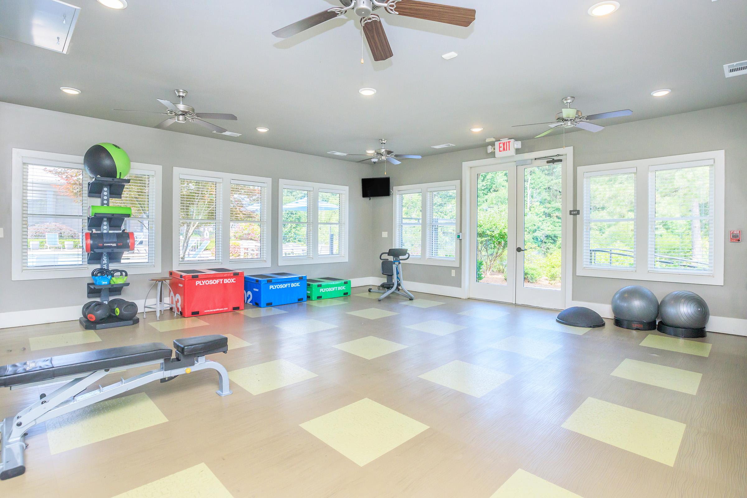A bright and spacious gym area featuring exercise equipment including benches, a weight rack, stability balls, and plyometric boxes. Large windows provide natural light and a view of the outdoors, creating an inviting atmosphere for workouts. The room has ceiling fans for ventilation.