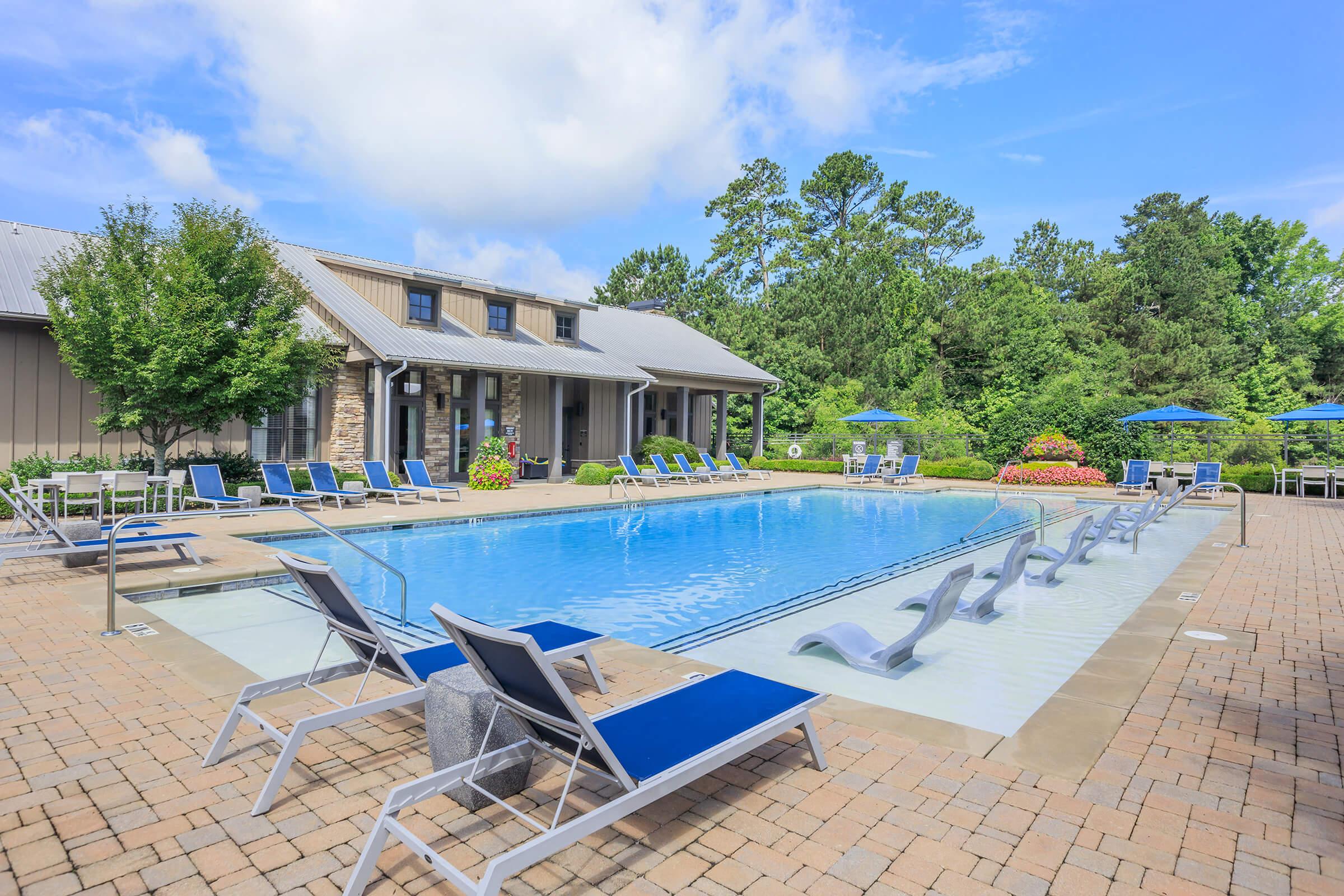 A sunny outdoor pool area featuring a sparkling blue pool surrounded by lounge chairs. Trees and colorful flower beds provide greenery in the background, and umbrellas offer shade. A building with a stone facade is visible near the pool, creating a relaxing atmosphere.