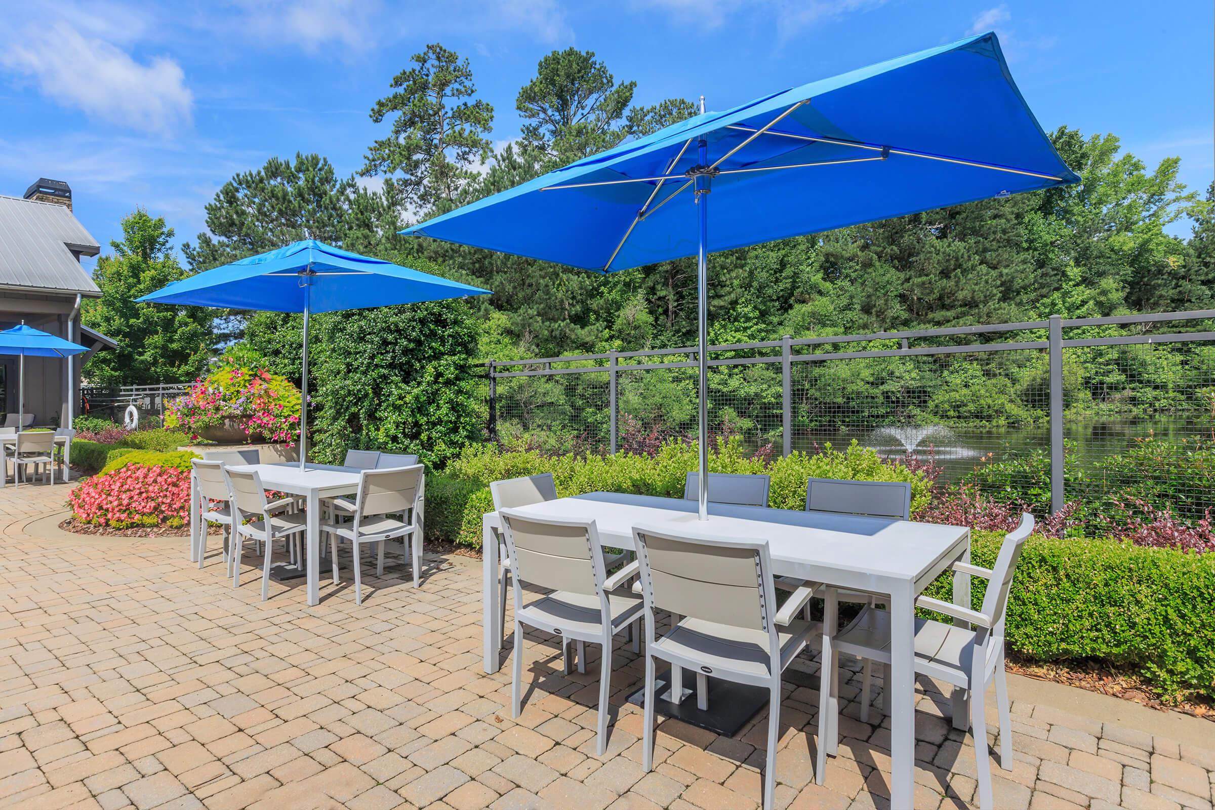 Outdoor seating area featuring two tables with light gray chairs under large blue umbrellas. The patio is paved with stone and surrounded by lush greenery and blooming flowers, creating a vibrant and relaxing atmosphere. In the background, there is a serene pond visible through a fence.