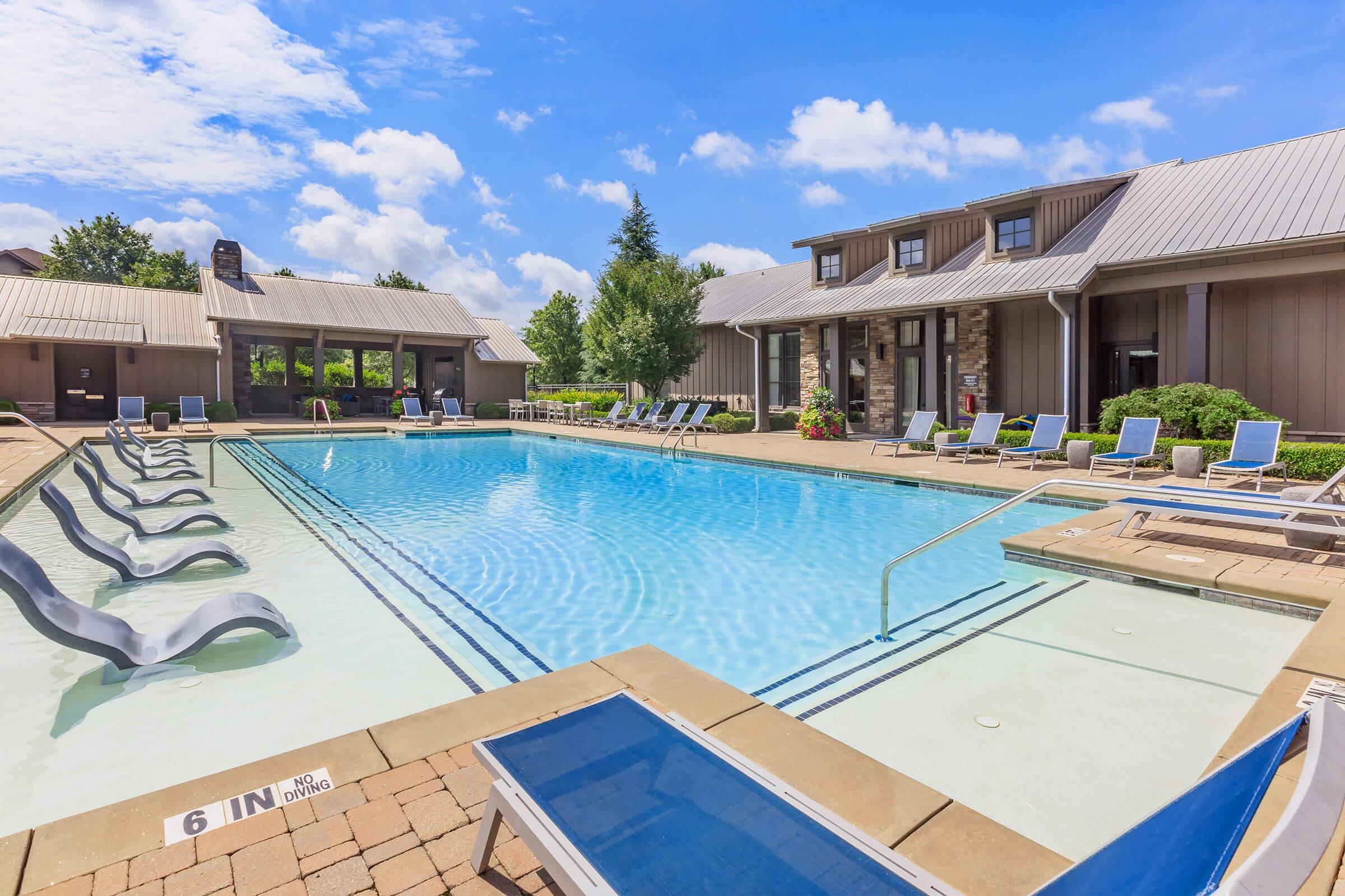 A spacious outdoor swimming pool surrounded by lounge chairs and a neatly landscaped area. The pool features a shallow end marked with "6 IN," and the backdrop includes cozy buildings under a bright blue sky with fluffy clouds.