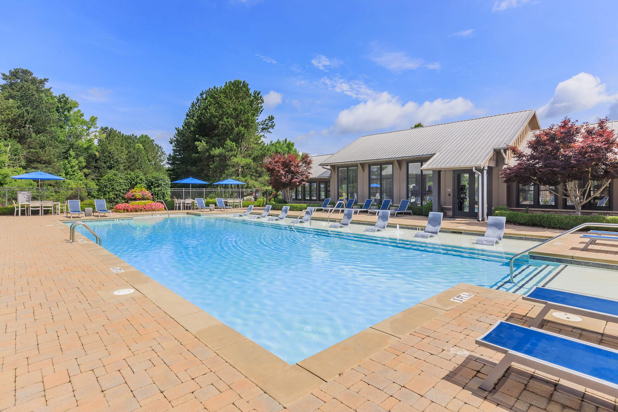 A bright, inviting outdoor swimming pool surrounded by lounge chairs and vibrant landscaping. The pool area features several umbrellas for shade and is located near a modern building. Lush greenery and colorful flowers add to the relaxing atmosphere under a clear blue sky.