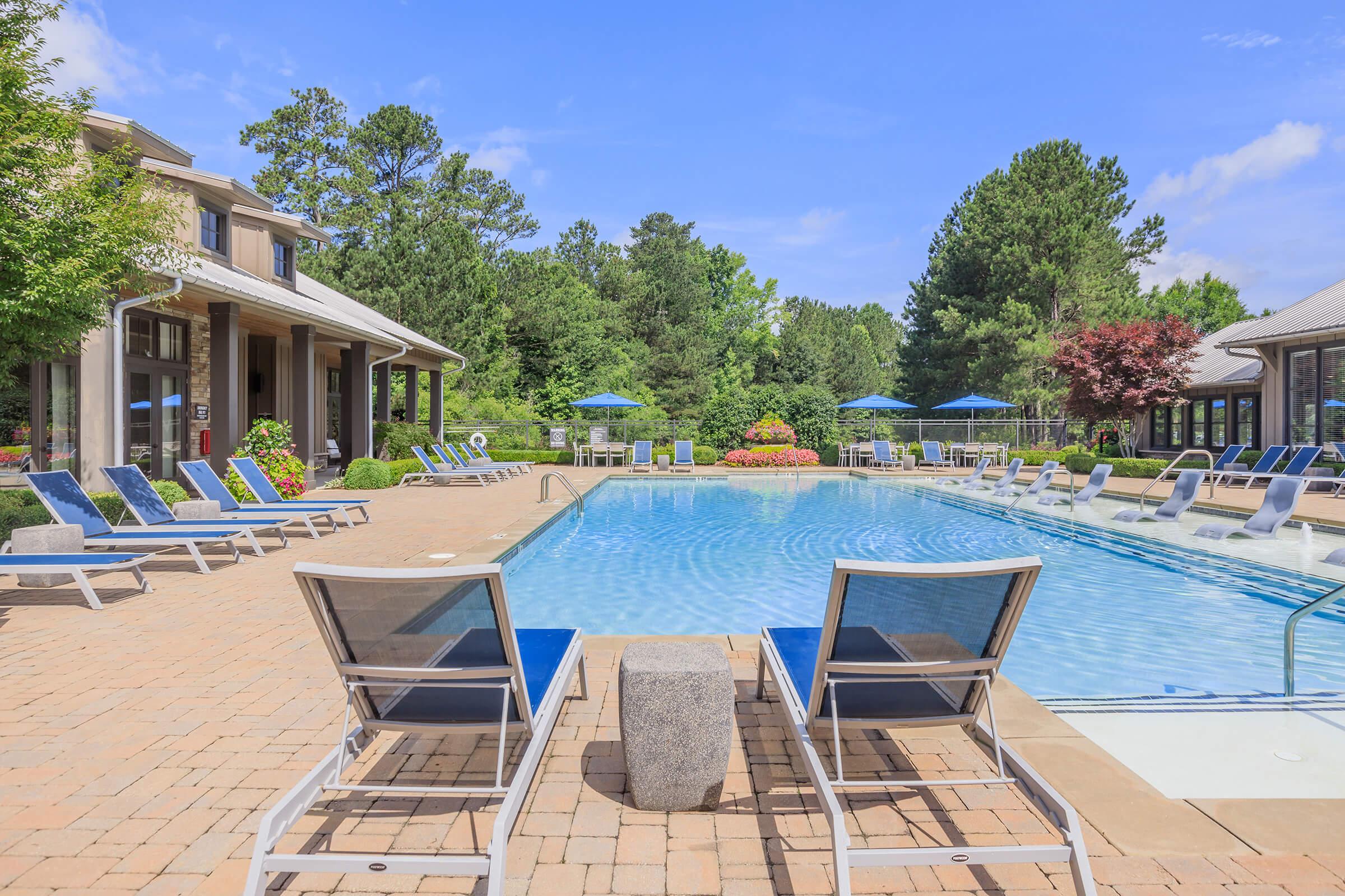 A serene outdoor pool area featuring multiple lounge chairs, surrounded by greenery and colorful flower beds. The pool is clear and inviting, with sun umbrellas in the background. A well-maintained building is visible on the left side, enhancing the relaxing atmosphere of the space.