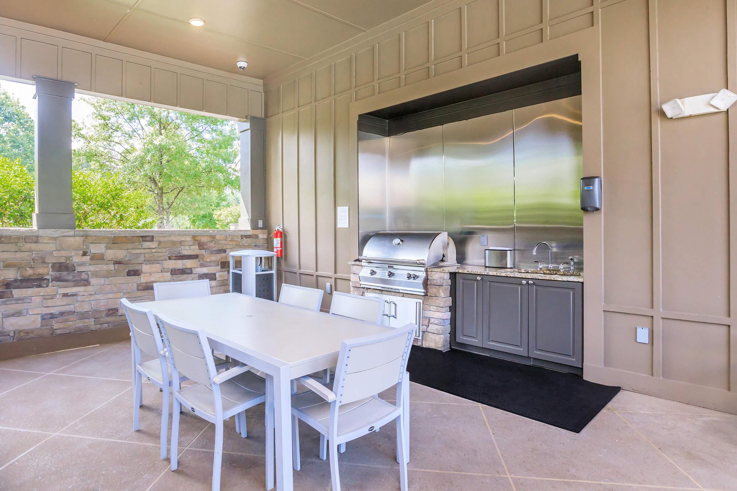 Outdoor kitchen area with a built-in stainless steel grill, sink, and cabinet storage. A white dining table with matching chairs is positioned nearby, set against a backdrop of stone walls and lush greenery. Natural light filters through the space, creating a welcoming atmosphere for outdoor dining and gatherings.