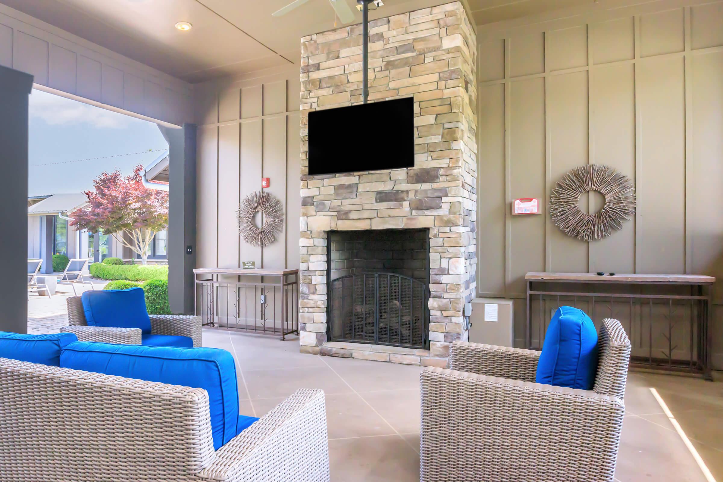 Cozy outdoor lounge area featuring a stone fireplace with a mounted TV above it, surrounded by two blue cushioned chairs and decorative wall accents. Natural light filters in, highlighting the neutral-toned walls and the inviting atmosphere.