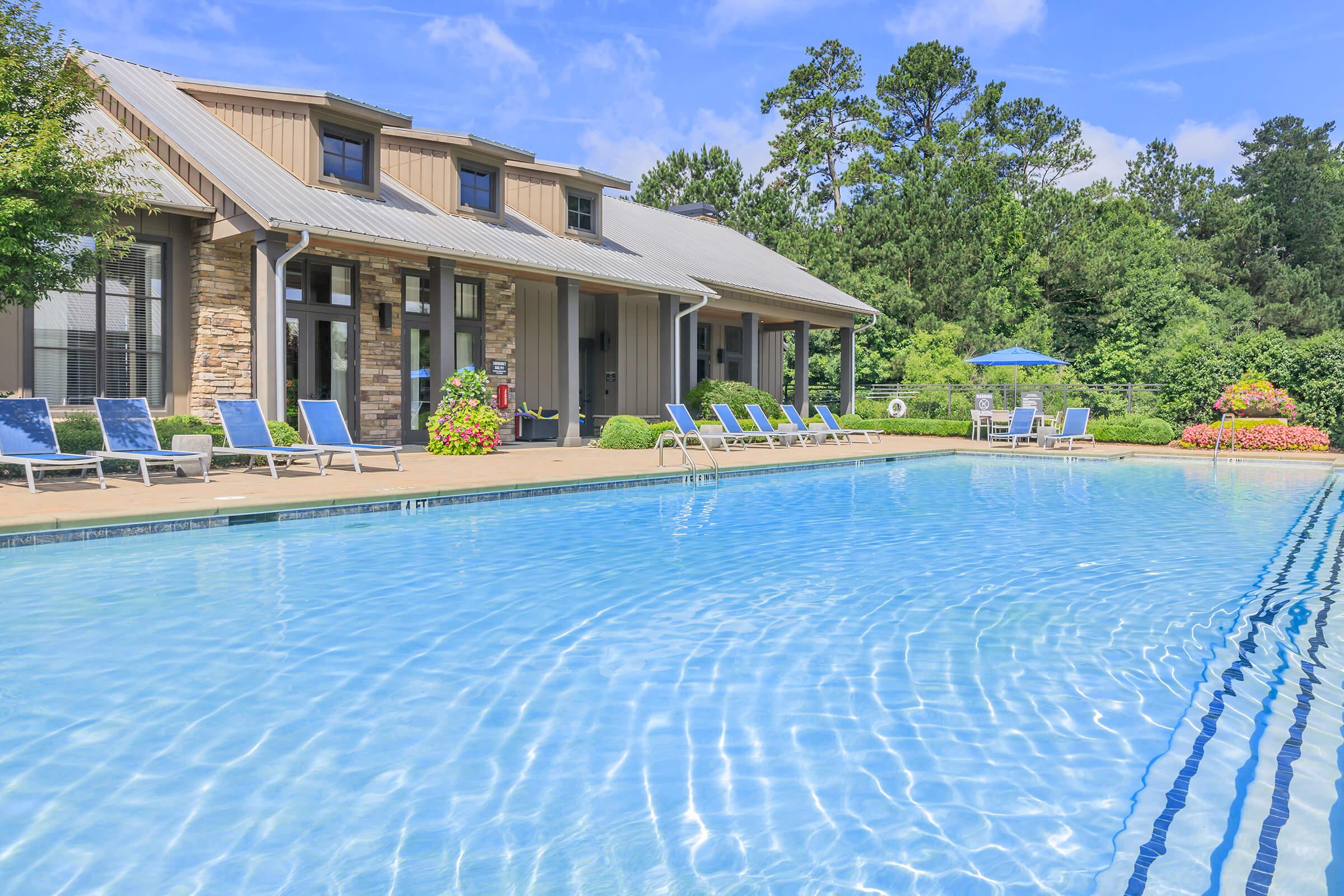 A clear blue swimming pool surrounded by lounge chairs, with a building featuring a stone facade in the background. Lush greenery and colorful flower beds enhance the scenery, under a bright blue sky with scattered clouds.